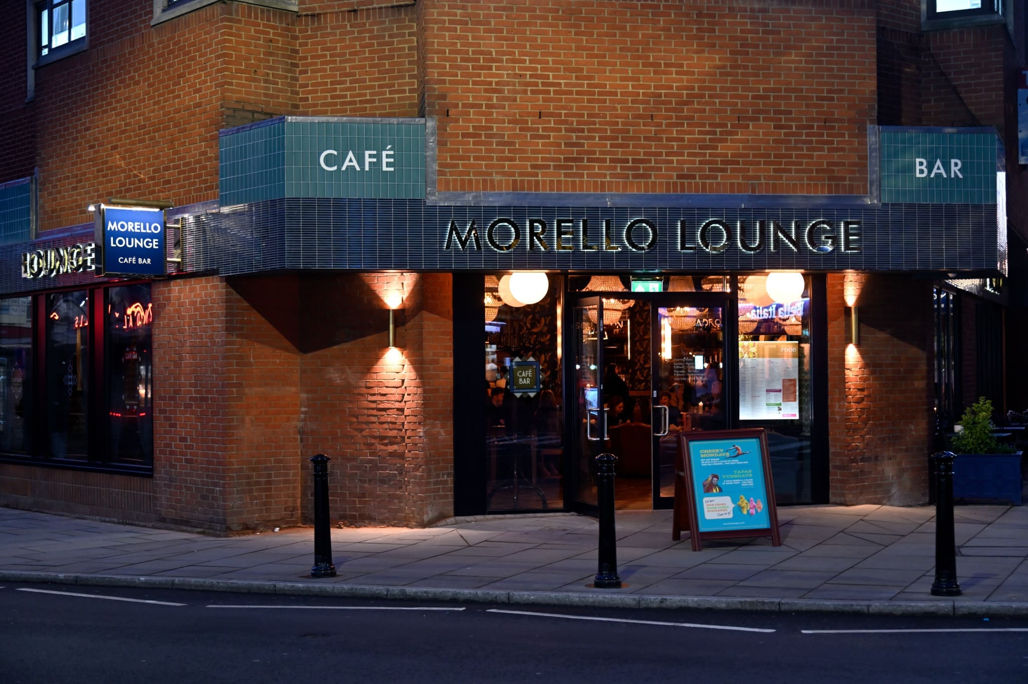 The image shows the exterior of Morello Lounge, a vibrant café bar on a street corner. Its warm lights illuminate the entrance, while a signboard outside invites guests into the stylish brick building with signature Morello blue-green tiled accents.