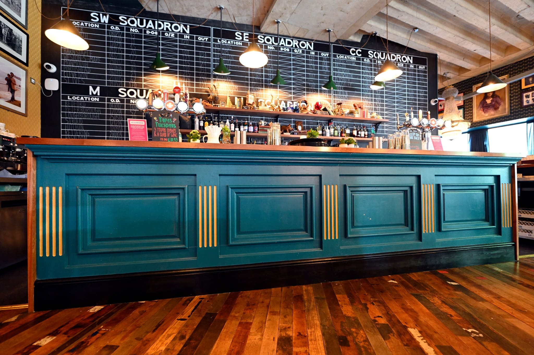 A stylish Morello bar with a teal counter, gold accents, and a wooden floor. Behind the bar are shelves with bottles, glasses, and decor, plus a large blackboard displaying drink options. Modern pendant lights hang above.
