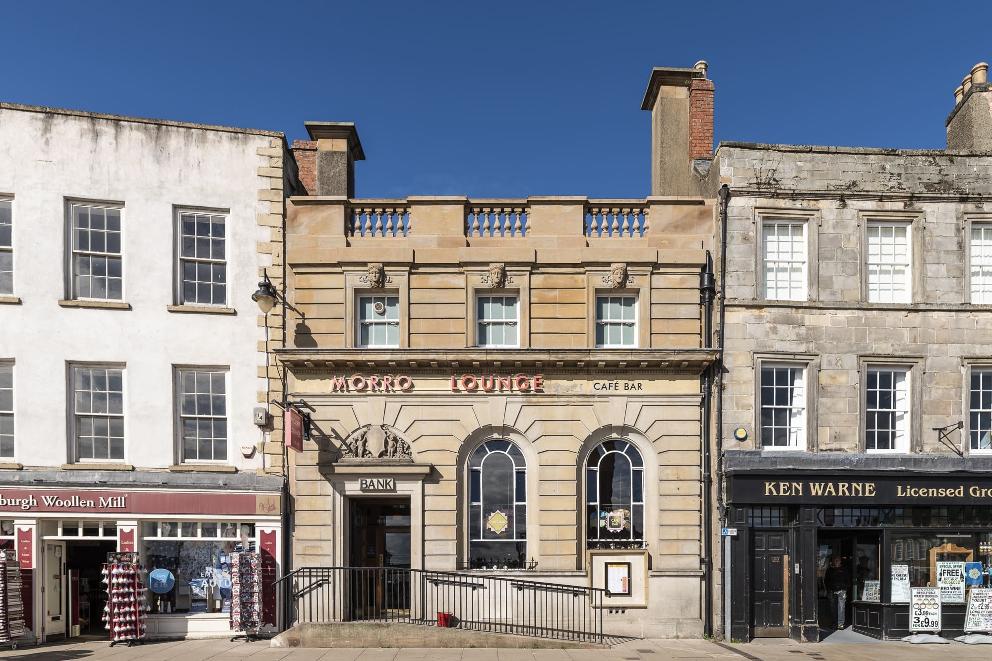 A stone building with arched windows houses Morro Lounge cafe bar, situated between a woolen mill shop and a licensed grocer on a sunny day under a clear blue sky.