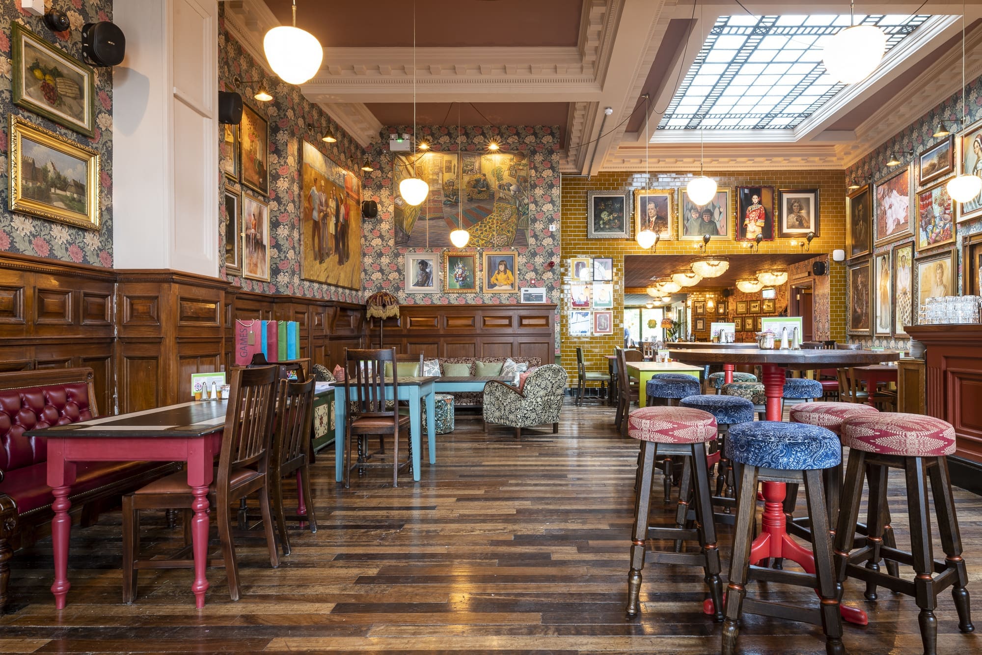 Eclectic Morro restaurant interior with wooden floors, colorful chairs, patterned wallpaper, framed artwork on the walls, hanging globe lights, and a large skylight above the bar area.