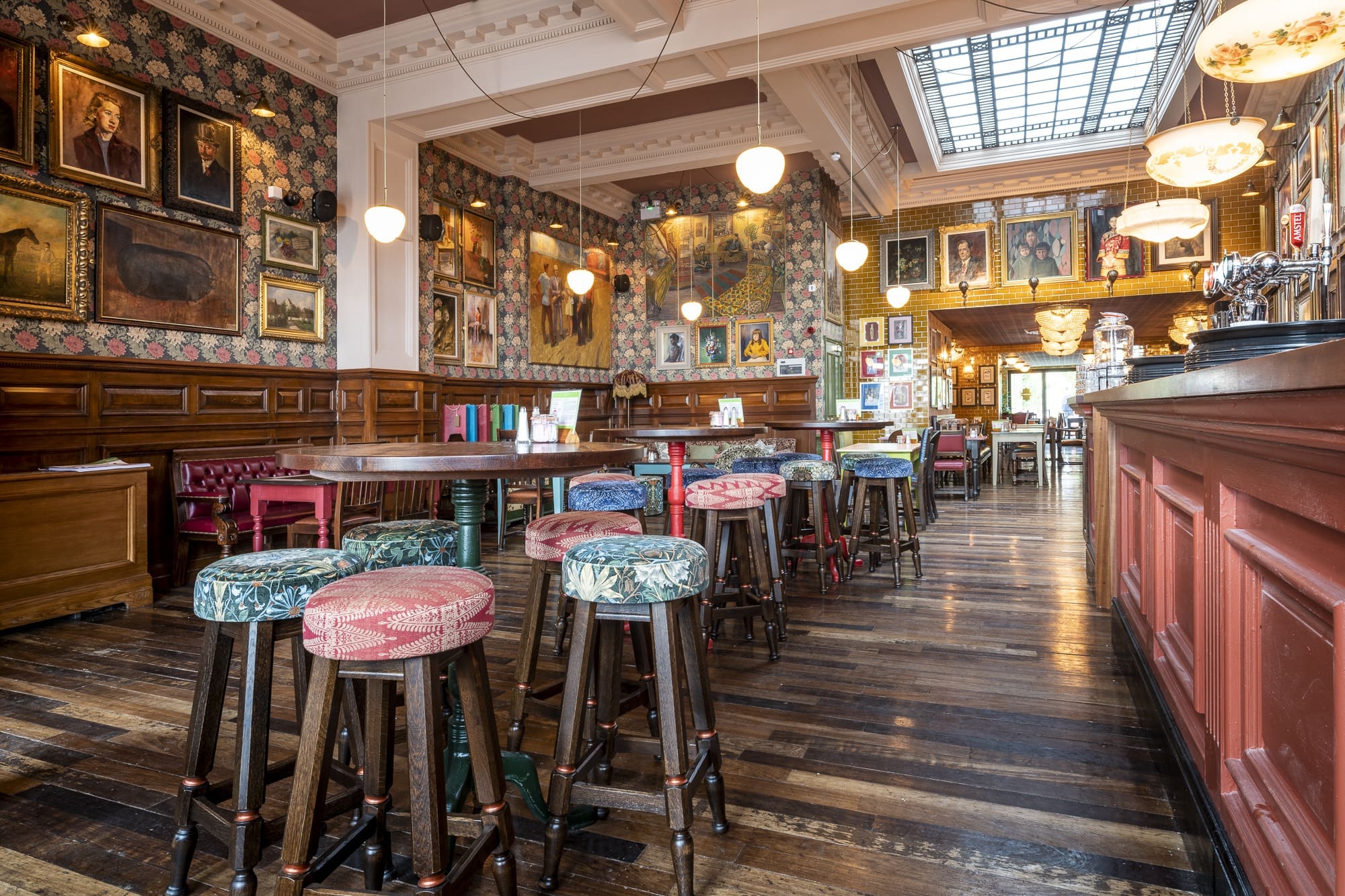 A cozy Morro pub interior with patterned wallpaper, vintage framed art, colorful cushioned bar stools, wooden floors, and warm lighting. The bar is on the right, and seating areas line the walls.