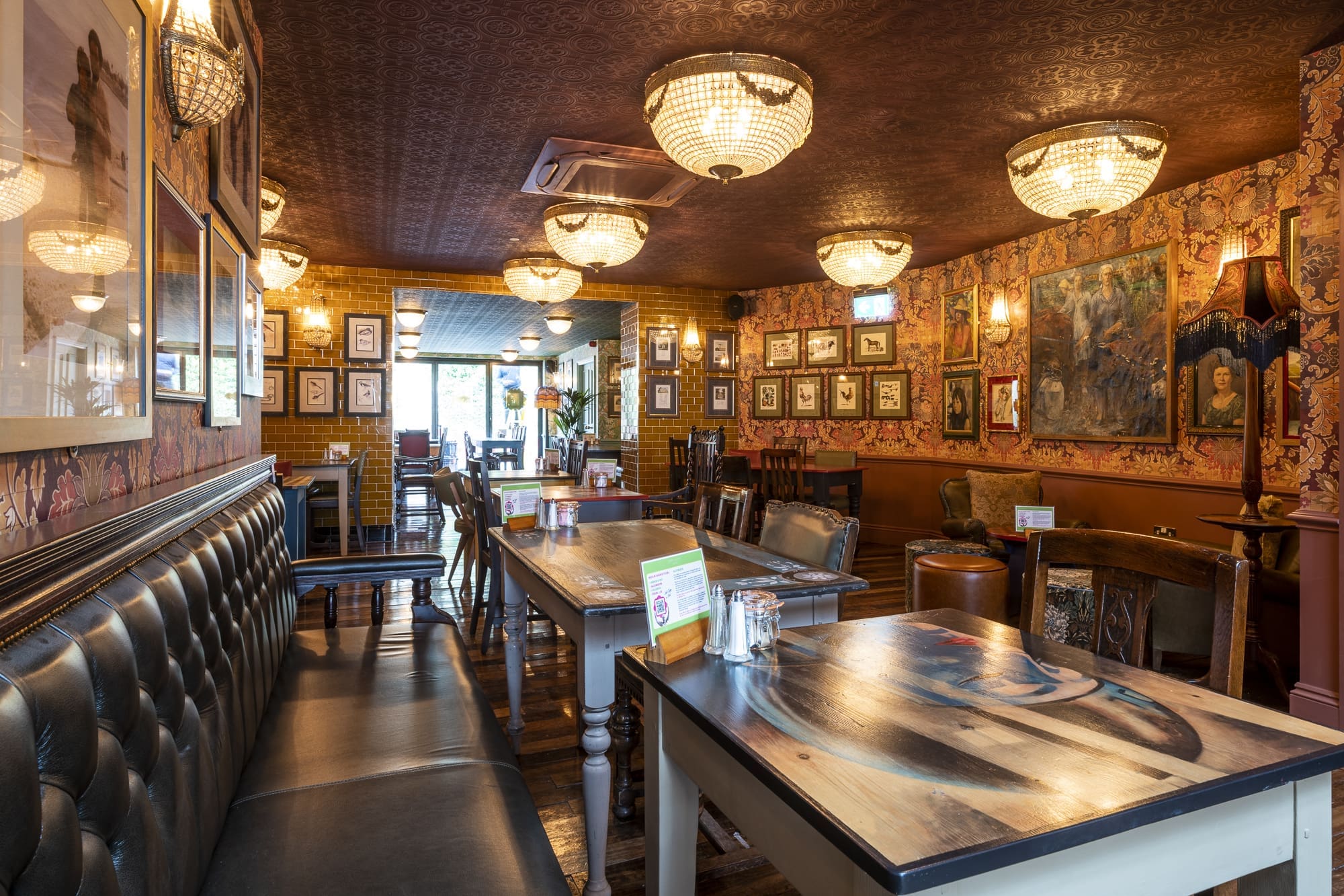A cozy, warmly lit Morro pub interior with leather benches, wooden tables, vintage chandeliers, patterned wallpaper, and framed art on the walls. The room is empty and inviting, with sunlight streaming through the windows.