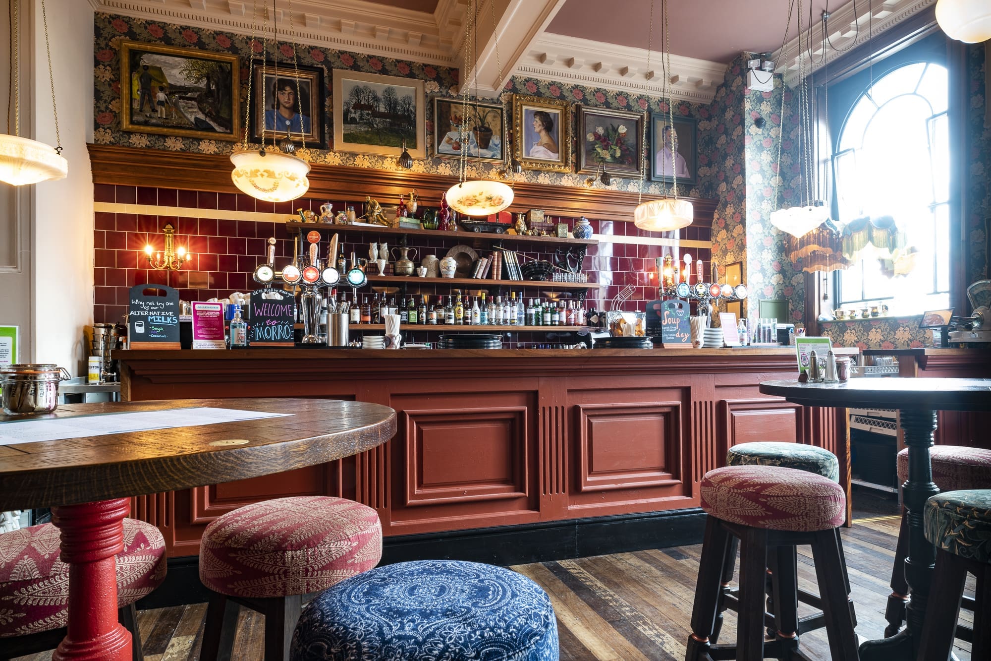 A cozy pub interior with patterned stools, round wooden tables, and ornate Morro artwork above a red paneled bar. Pendant lights hang from the ceiling, while bottles and taps are neatly arranged behind the bar.