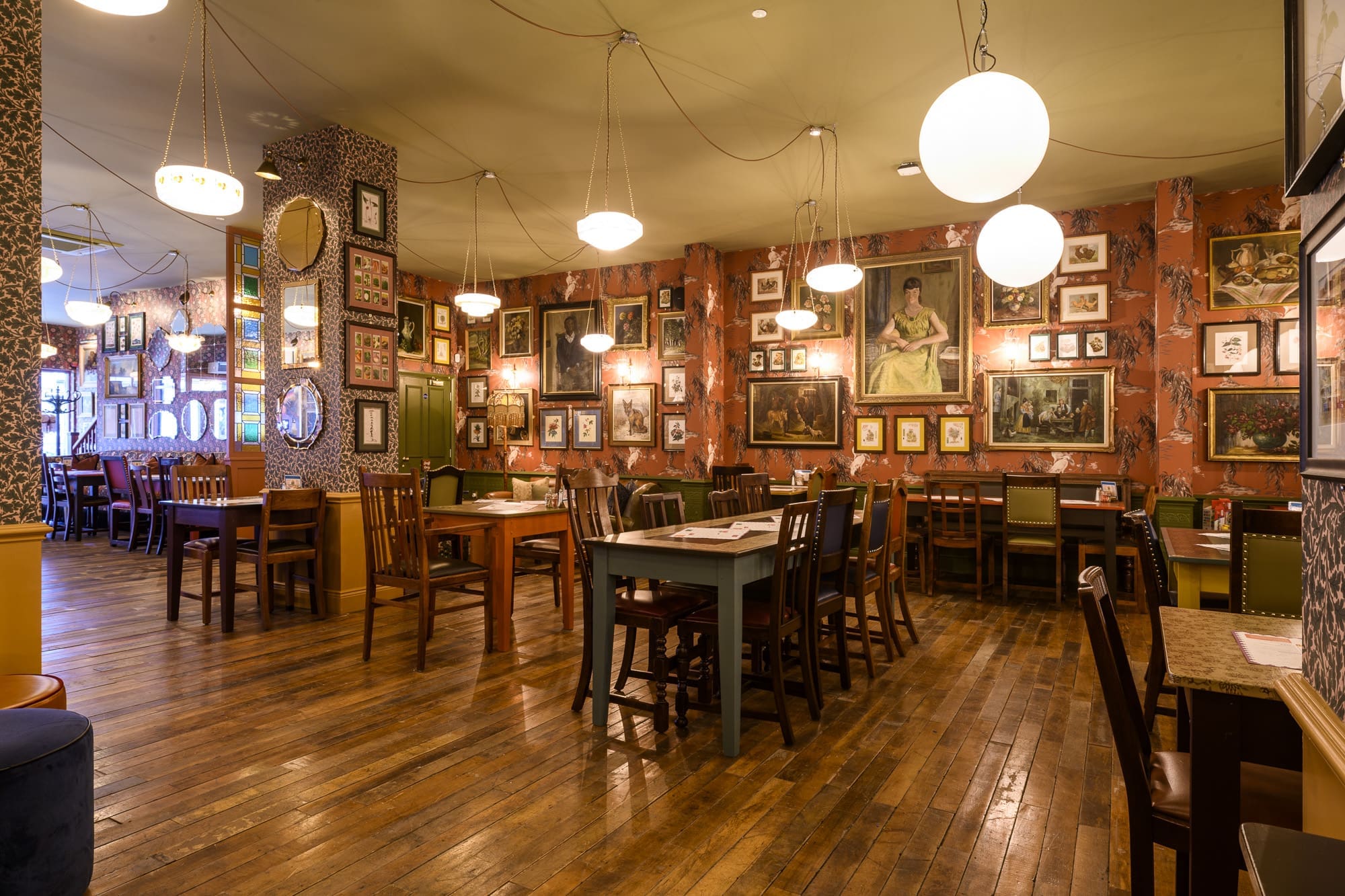 A cozy restaurant interior with wooden floors, mismatched tables and chairs, round hanging lights, and a muro covered in framed photos and paintings. The space is empty and warmly lit.