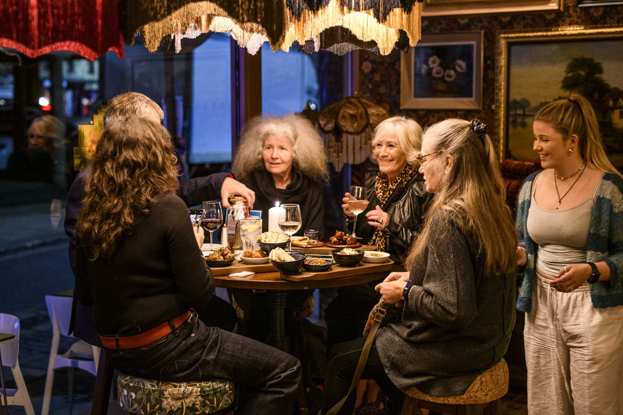 A group of older women sit around a table in a cozy, warmly lit restaurant, sharing food, drinks, and laughter. A younger woman stands nearby, smiling and engaging with the group. Paintings adorn the muro behind them as lamps cast a gentle glow.