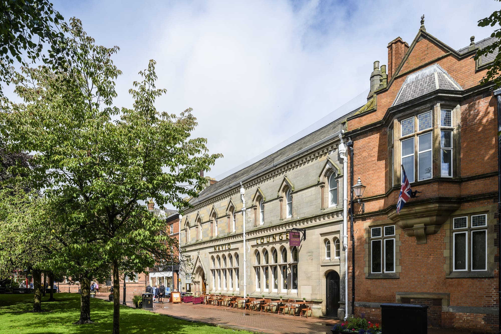 A historic stone and brick building with large windows and outdoor seating near Navio is bordered by a tree-lined walkway and green grassy area on a bright, partly cloudy day.