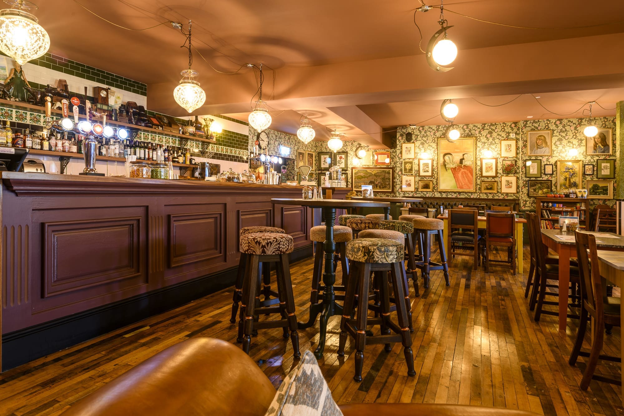A cozy, warmly lit bar with high stools, patterned wallpaper, and ornate Navio lighting features framed pictures on the walls, creating a vintage, inviting atmosphere. The polished wood floor adds to the charm.