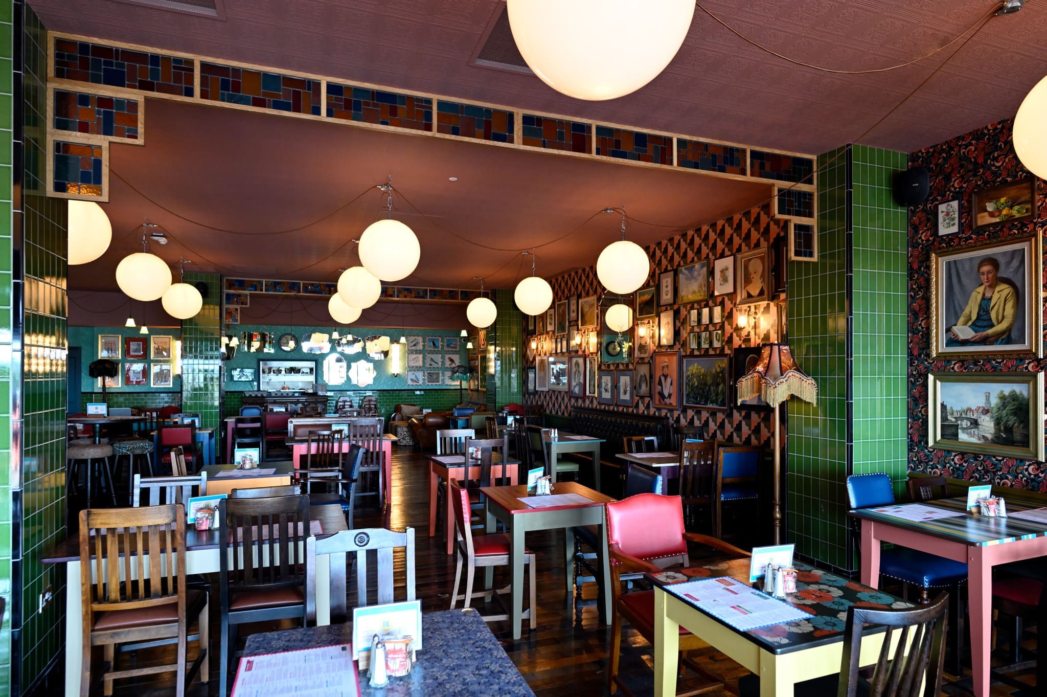 Colorful, eclectic restaurant interior with mismatched chairs, patterned walls, round hanging lights, and framed art on the walls. Novo’s cozy atmosphere invites you to relax as you browse menus at vibrantly set tables.