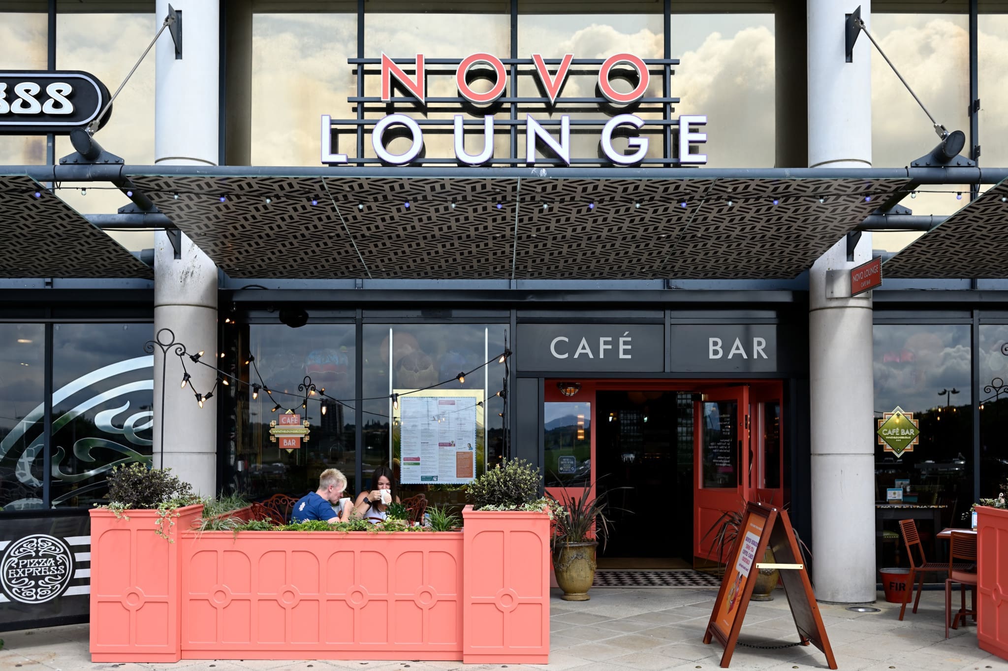 Two people sit at a pink outdoor table in front of Novo, the café and bar with large glass windows, a patterned awning, string lights, and visible entrance signs beside the menu board.
