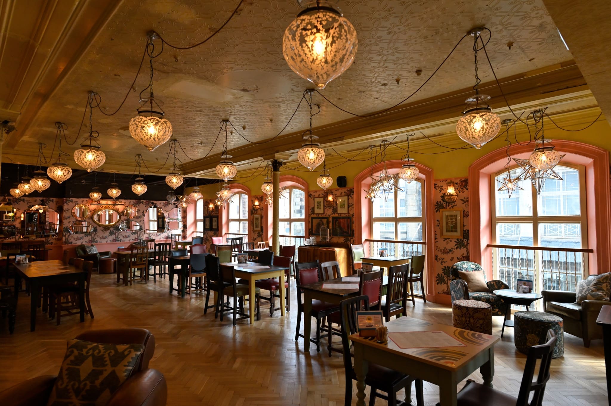 A cozy, warmly lit café with Occulo-inspired hanging lanterns, wooden tables and chairs, colorful walls, large windows, and eclectic decor creates an inviting and relaxed atmosphere.