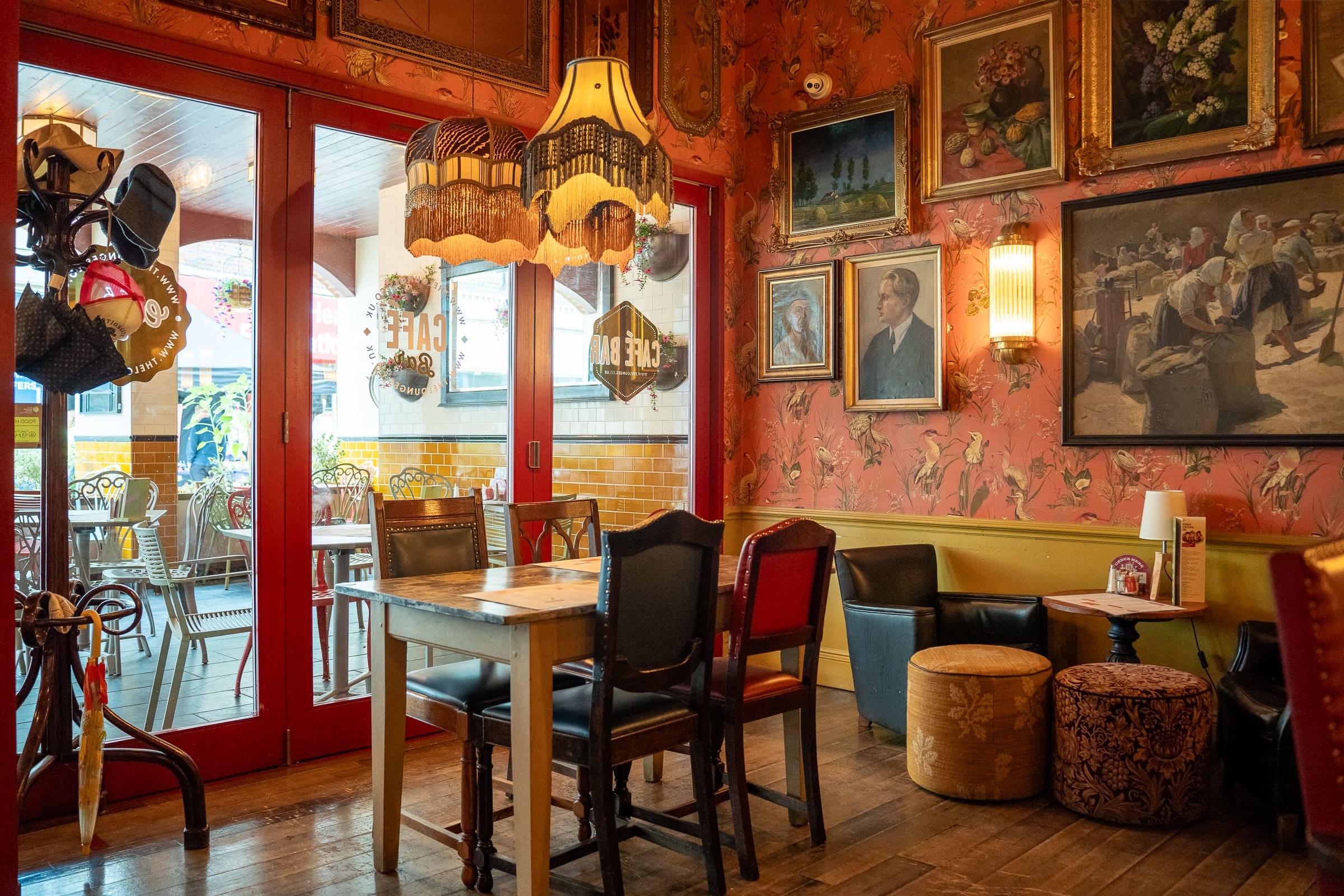 A cozy vintage café interior by Olmo with patterned wallpaper, framed paintings, eclectic chairs, and a fringed lamp above a wooden table. Through glass doors, more seating and natural light are visible outside.