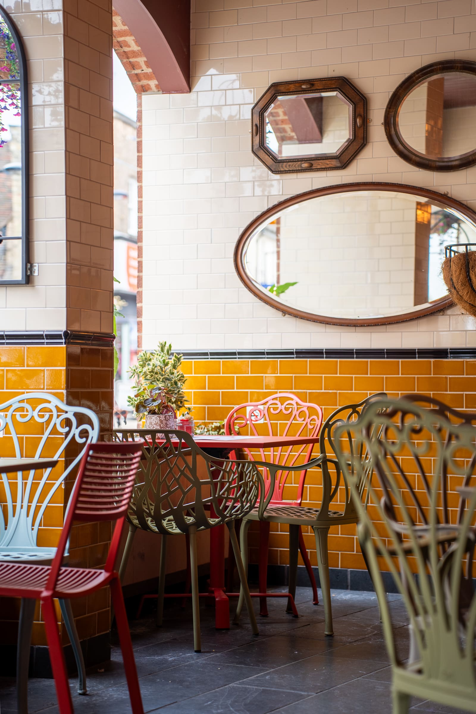 A cozy café corner by Olmo with colorful metal chairs, yellow-tiled walls, and three decorative mirrors reflecting light. A small plant sits on the round table by the window.