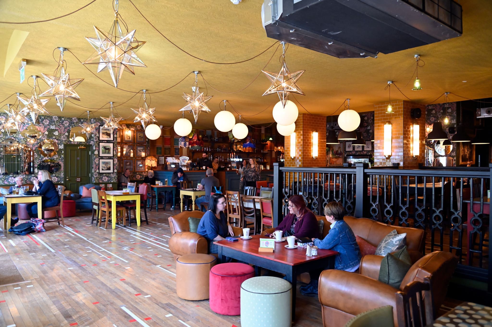 A spacious, eclectic cafe at Ostello with star-shaped and round hanging lights, colorful furniture, wooden floors, and groups of people sitting at tables and sofas, engaged in conversation and drinks.