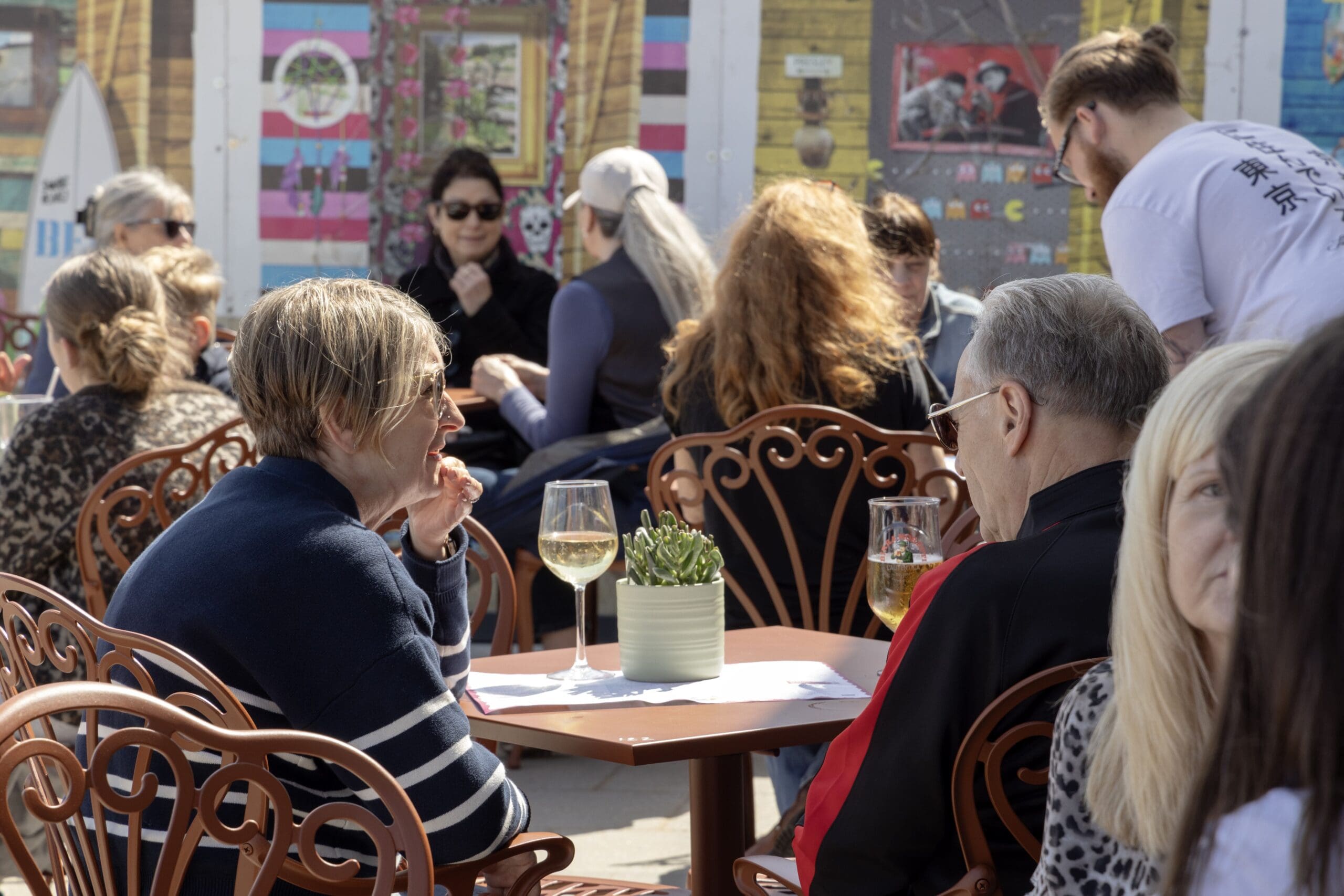 People sit at outdoor café tables, talking and enjoying drinks on a sunny day. A potted plant and a perico add vibrant life to the scene, with colorful murals and more people in the background for a lively, relaxed atmosphere.