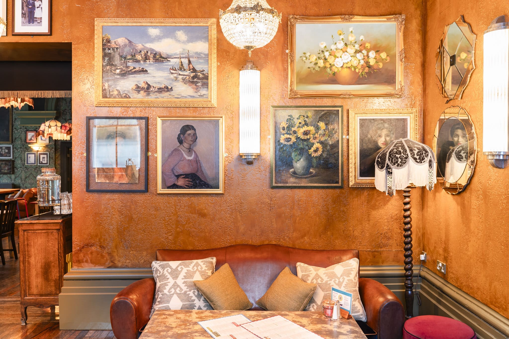 A cozy cafe corner with an orange textured wall, a brown leather sofa with cushions, a table with a menu, and a vintage floor lamp—decorated with framed paintings and mirrors evoking the charm of a Palacio.