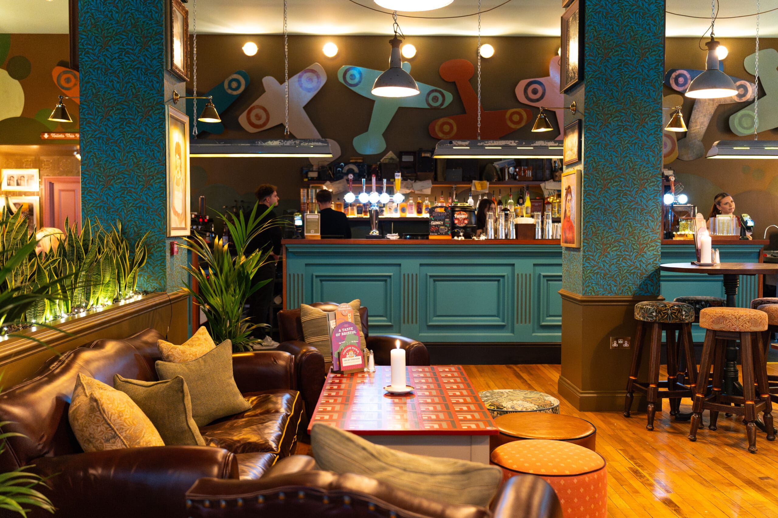 A cozy, colorful bar interior with patterned walls, leather sofas, cushions, plants, and a candle-lit coffee table; the turquoise Palomo bar counter glows in the background with stools and drinks on display.