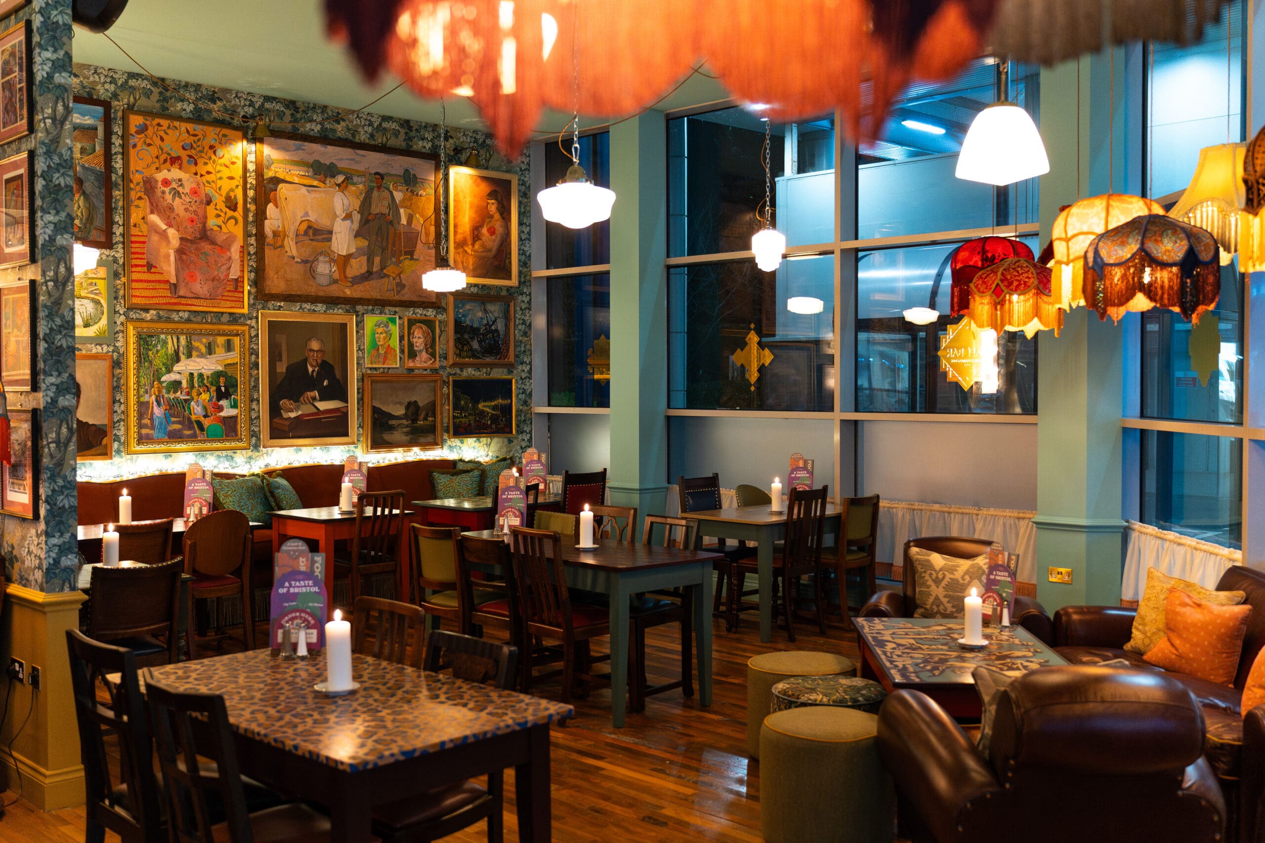 A cozy, warmly lit café with eclectic decor, featuring mismatched chairs, art-covered walls, patterned tables, cushioned seating, and various hanging lamps. Palomo-inspired touches and candlelit tables create an inviting atmosphere.