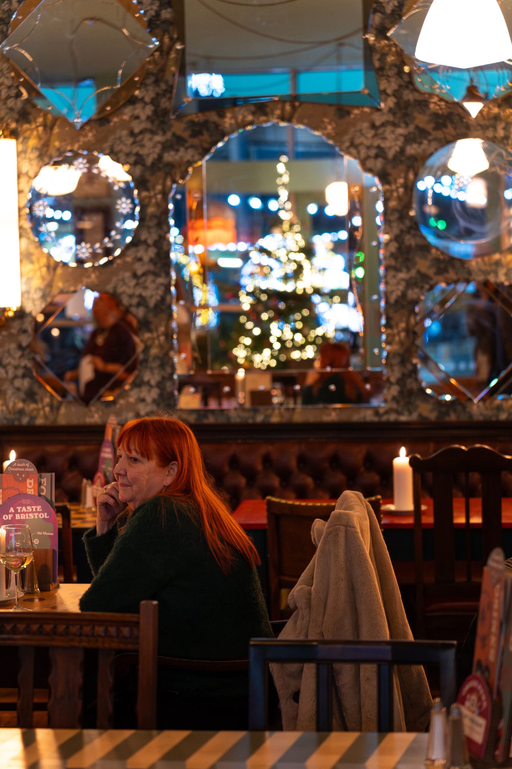 A woman with red hair sits alone at a lounge restaurant table, speaking on the phone. Behind her, a decorated Christmas tree and festive lights are reflected in circular mirrors, creating a warm, cozy atmosphere.