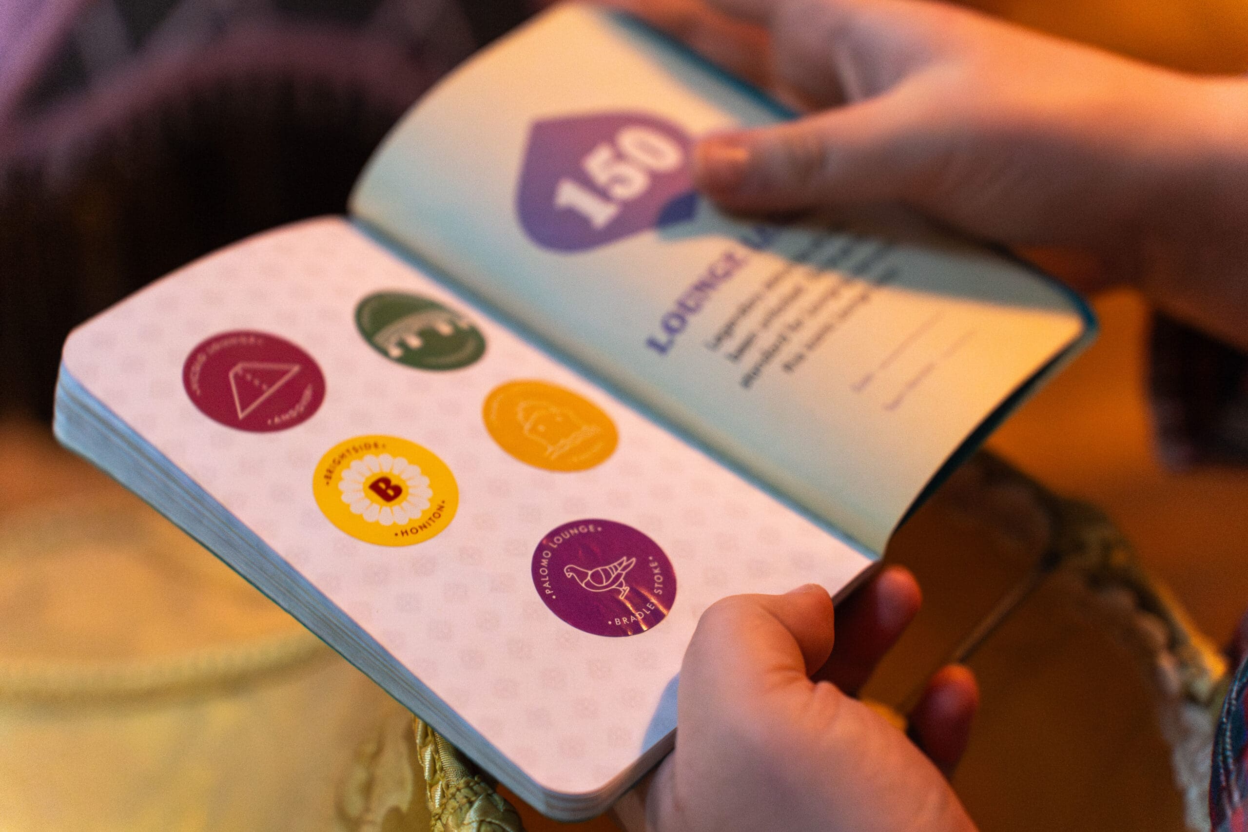 A person holds open a book featuring colorful palomo badges or stickers, each with unique symbols and numbers, on one page and text with a large "150" on the opposite page.