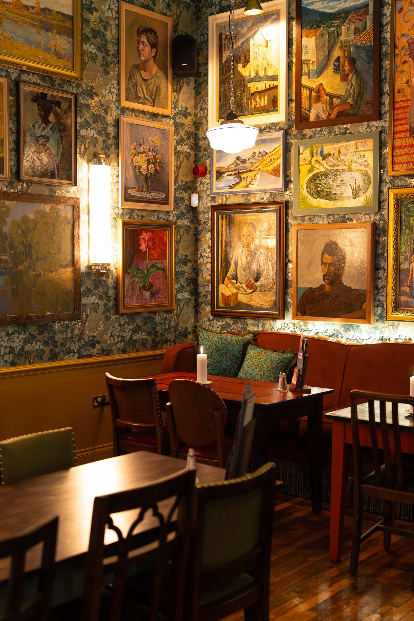 A cozy, warmly lit café with wooden tables and chairs, orange cushioned benches, and walls decorated with numerous colorful paintings and portraits in ornate frames, including a striking Palomo piece.