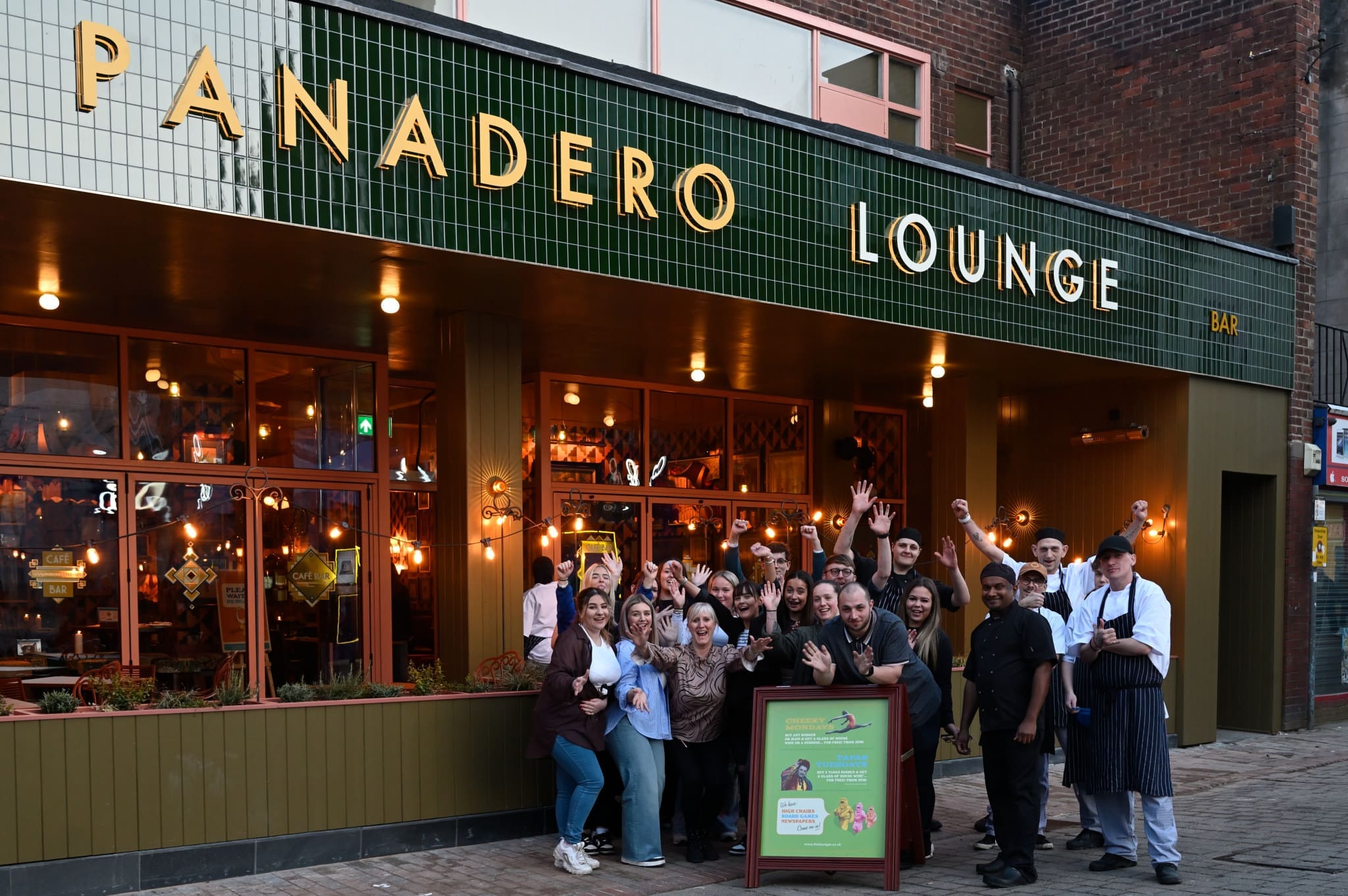 A group of people, likely panadero staff, pose and smile together outside Panadero Lounge bar and restaurant, which has green-tiled signage and warm lights glowing inside.
