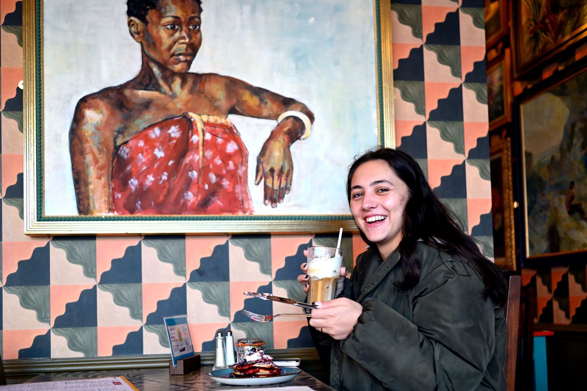 A smiling person with long dark hair holds a drink and a plate of food at a panadero’s table in a restaurant, with a large, colorful portrait of a woman in the background on the patterned wall.