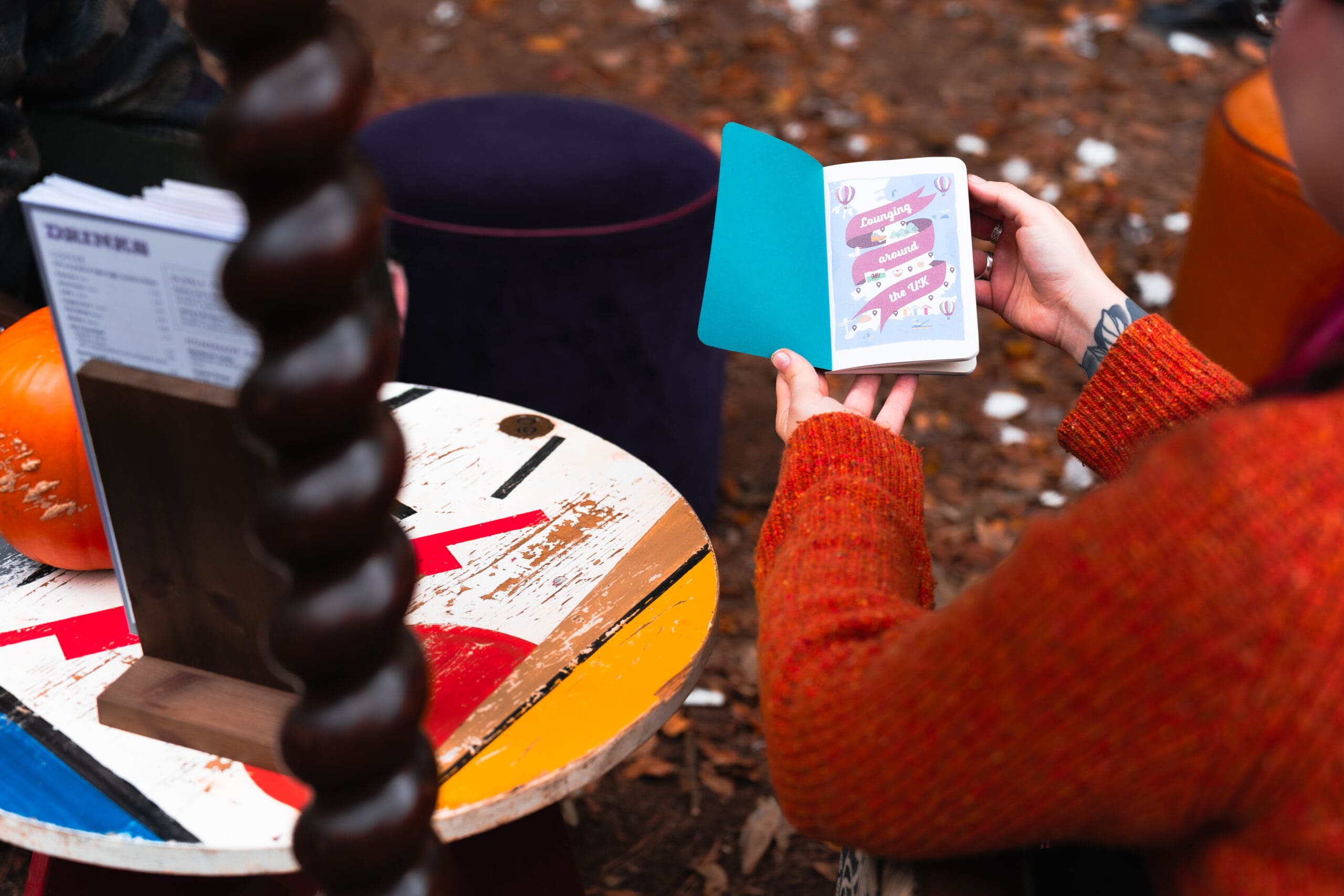 A person in an orange sweater lounges outdoors, holding an open greeting card next to a small, colorful table and a pumpkin, surrounded by autumn leaves.