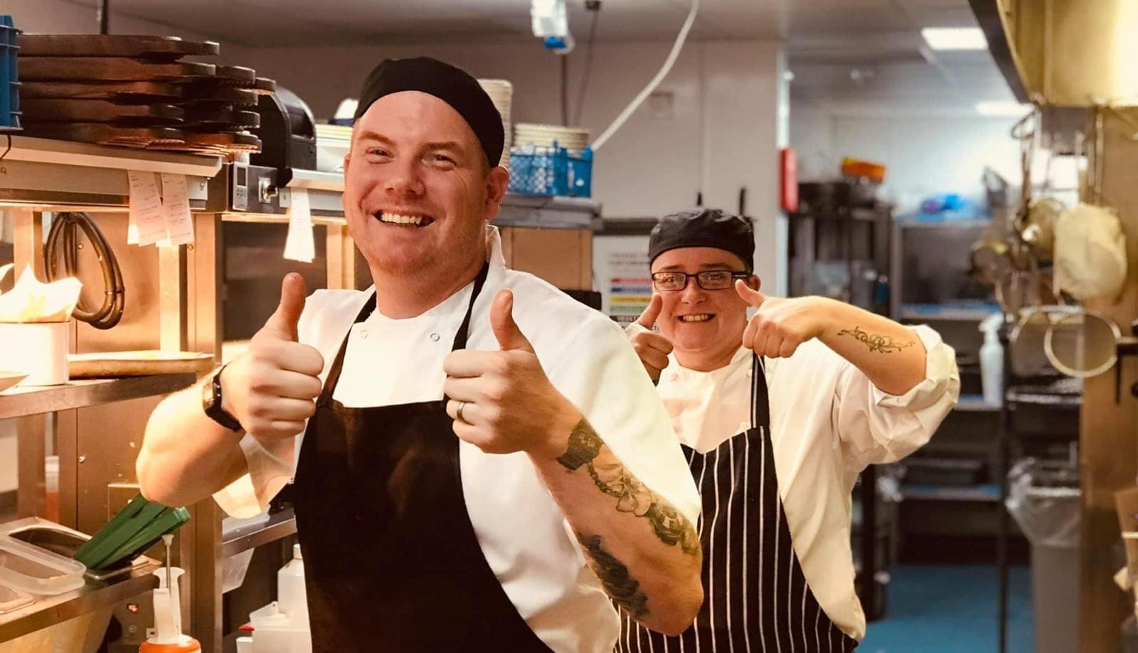 Two chefs in a kitchen, both smiling and giving thumbs up. They are wearing white chef coats, striped aprons, and black hats, standing among shelves and kitchen equipment. The background shows a busy kitchen environment.
