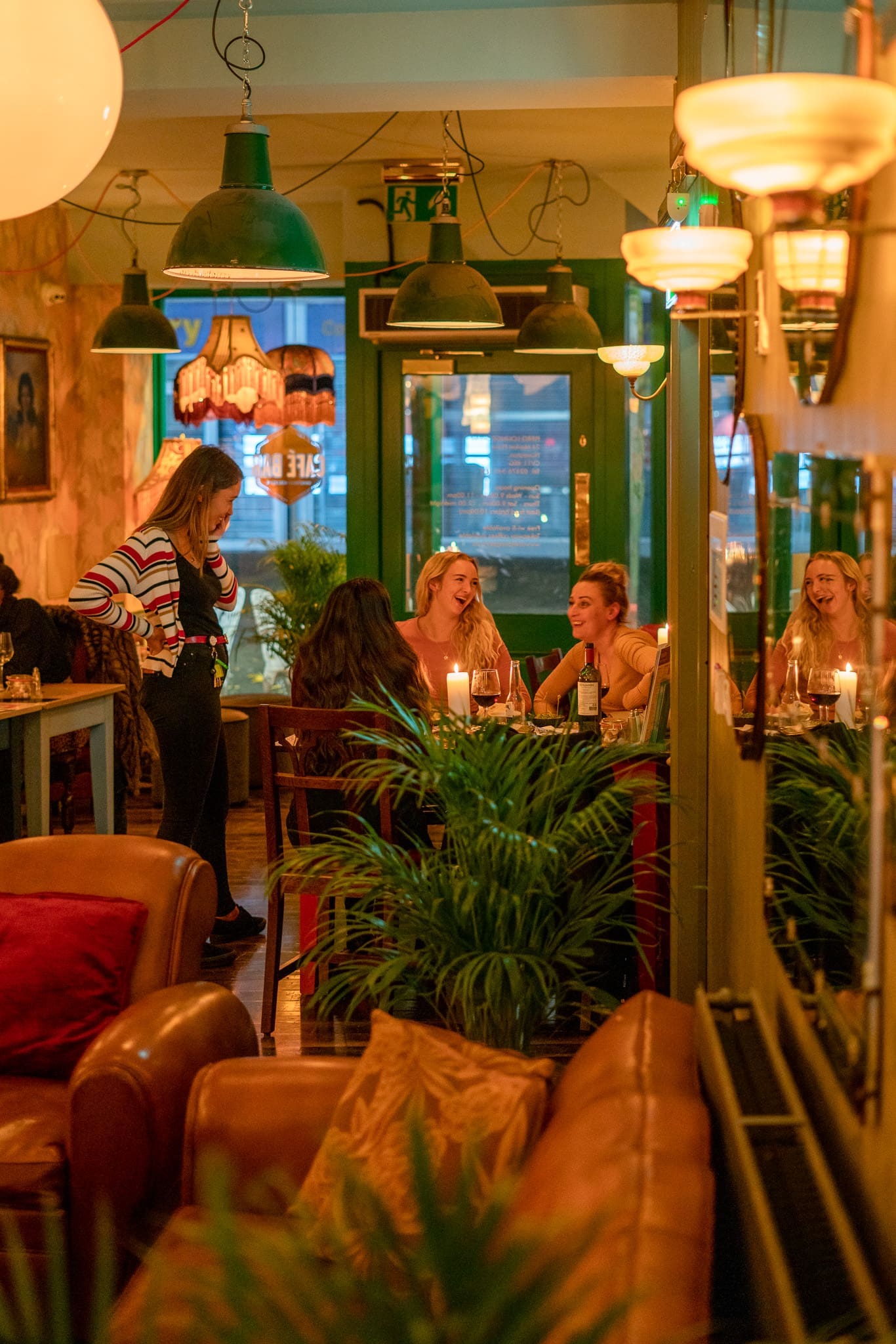 A cozy, warmly lit café with several women sitting at a table, laughing and talking by candlelight. One woman stands nearby. Piero’s touch is seen in the green plants, hanging lights, and cozy chairs that create a relaxed, inviting atmosphere.