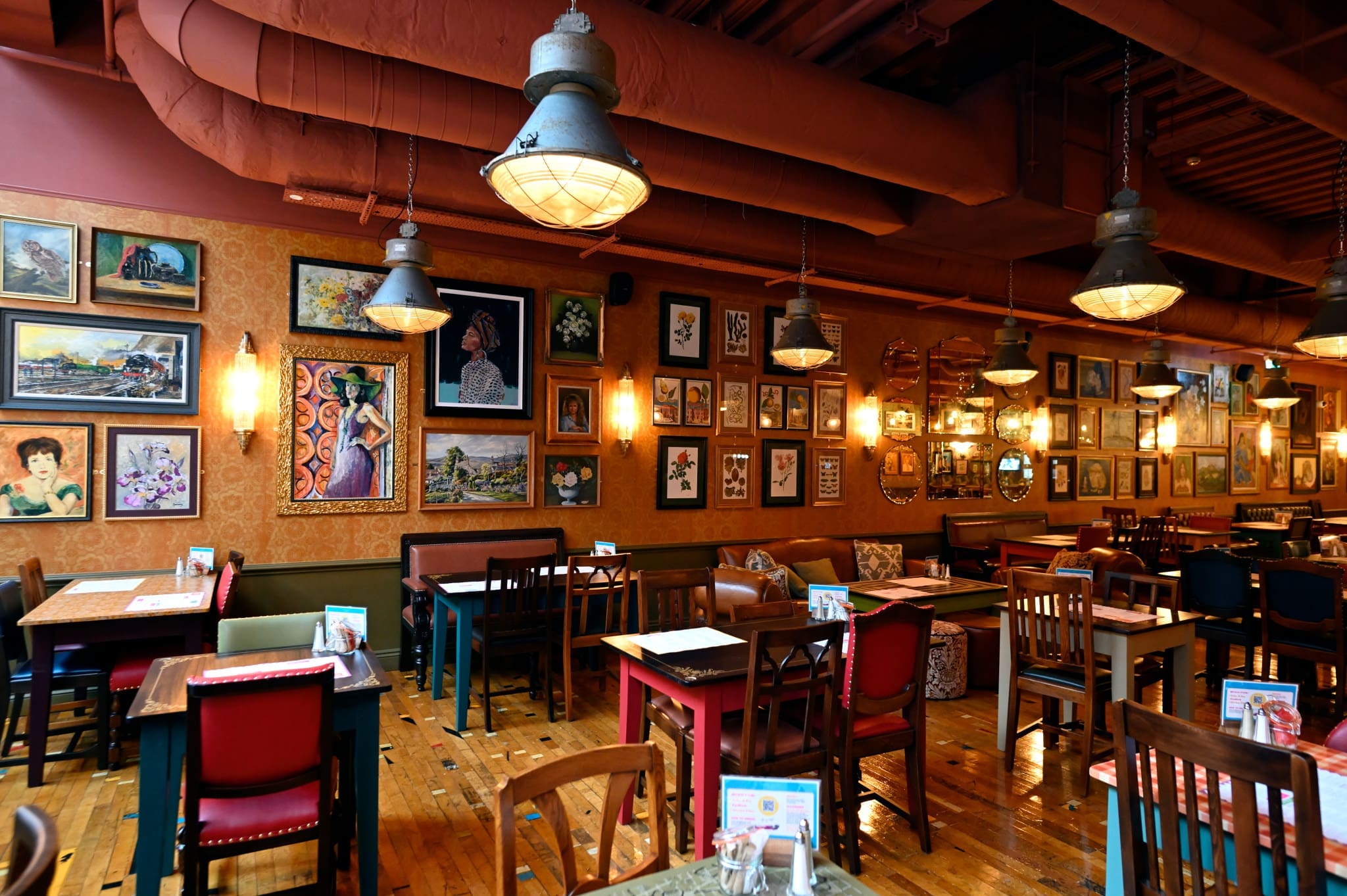 Cozy restaurant interior with wooden tables and mismatched chairs, framed artwork and paintings covering the walls, warm lighting from vintage lamps, and a polished pineto wooden floor. No people are present.