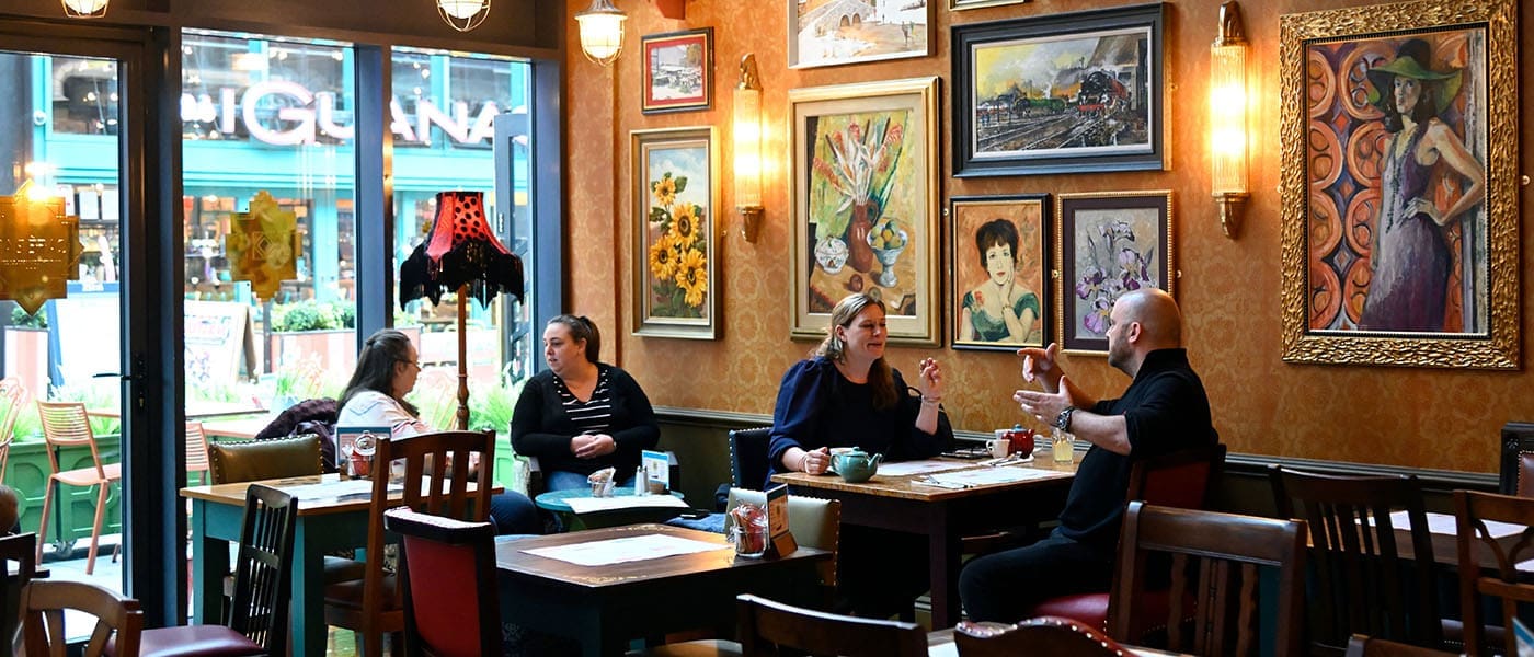 Two pairs of people sit at separate tables inside Pineto, a cozy, warmly lit café decorated with colorful paintings, vintage wallpaper, and wooden furniture. Large windows reveal a lively street scene outside.