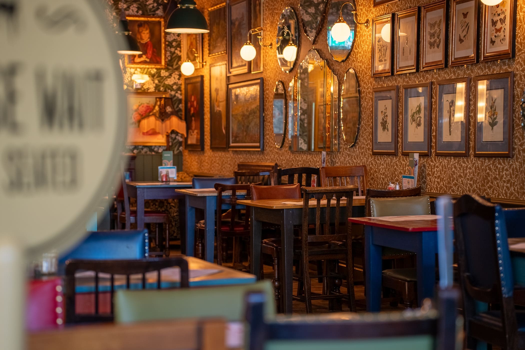 A cozy restaurant interior with colorful wooden tables and chairs, eclectic framed art on patterned walls, and warm hanging lights creates a welcoming Pinto-inspired atmosphere.