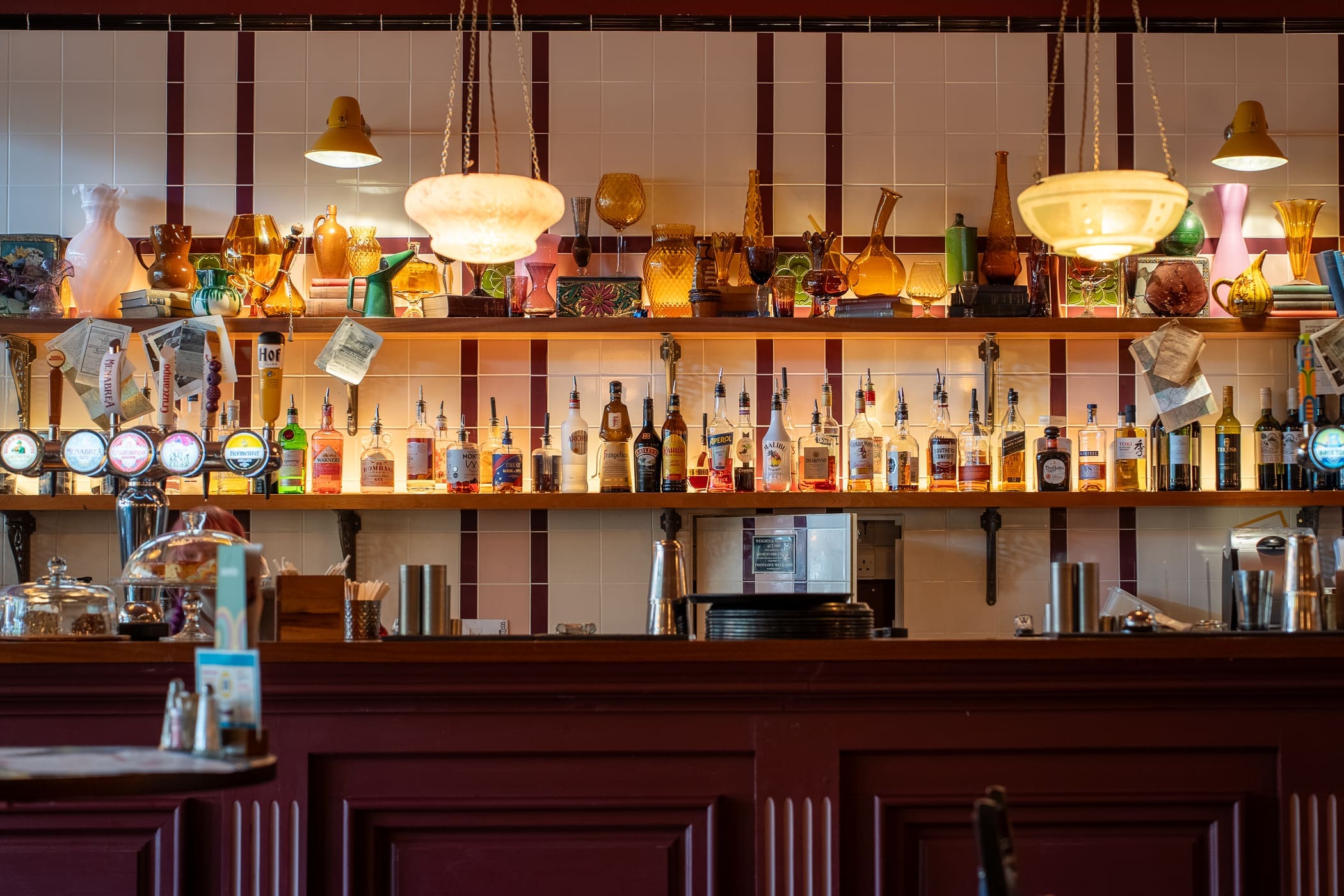 A bar with Pinto shelves displaying various bottles, glassware, and beer taps. Warm pendant lights hang above, illuminating the countertop and eclectic decorations against a tiled wall.