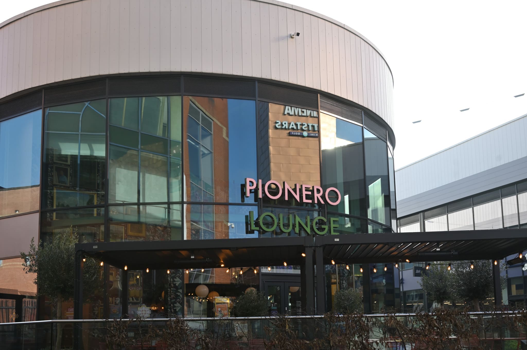 Exterior view of Pionero Lounge, a modern glass-fronted pionero building with neon signage, outdoor seating area, string lights, and plants, featuring reflections of nearby buildings in the large windows.