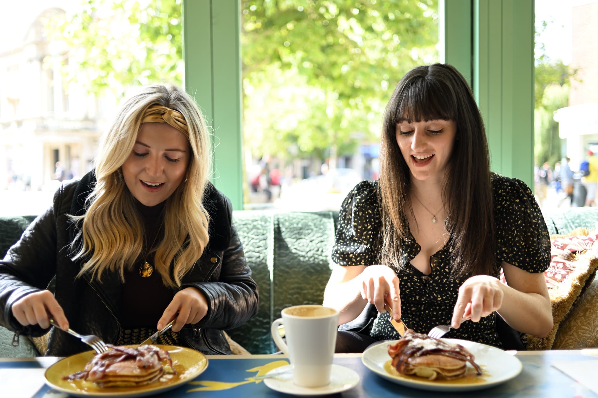 Two women sit at a sunlit café table at Ponto, smiling and cutting into pancakes topped with bacon. A cup of coffee sits between them, while large green-framed windows reveal a bright, leafy street outside.