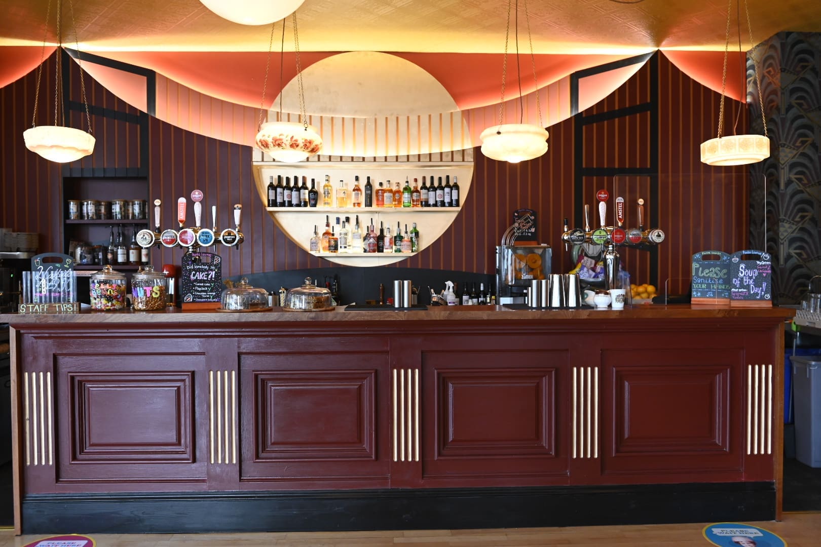 Ponto is a stylish bar with a red and gold art deco theme, featuring a wide wooden counter, hanging globe lights, beer taps, and colorful chalkboard signs on each end of the bar.