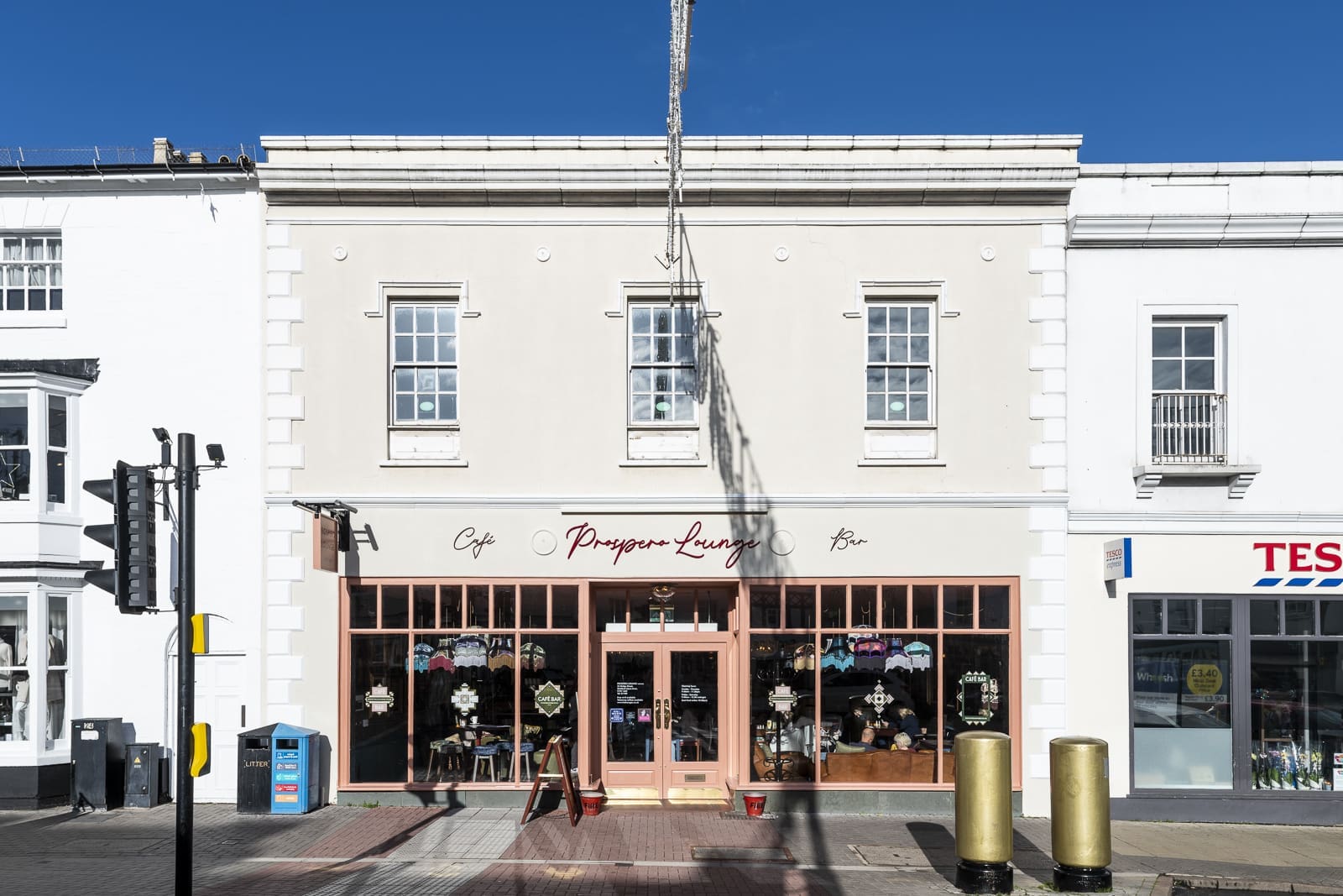 A light-colored café and bar named "Prosper Lounge," inspired by Prospero, with large front windows and peach trim, is situated between other shops on a sunny street. A Tesco store is visible on the right.