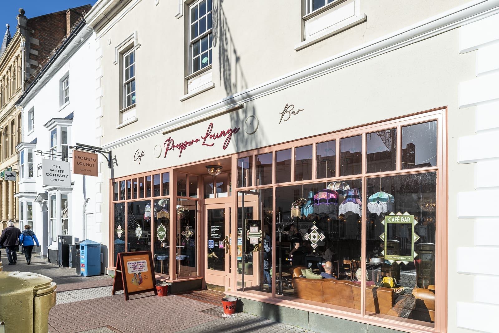 Street view of Prospero’s café and bar, "Prosecco Lounge," with large windows, pink frames, and outdoor signage. People walk by on the sidewalk or relax inside the brightly lit café.