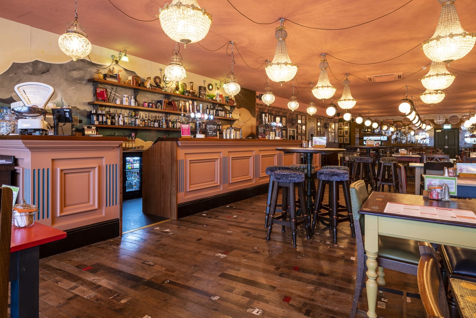 A cozy bar with peach-colored counters, wooden stools, and vintage chandeliers hanging from a salmon-pink ceiling. Shelves with bottles and glasses line the wall, creating a Prospero-inspired atmosphere among mismatched tables and chairs.