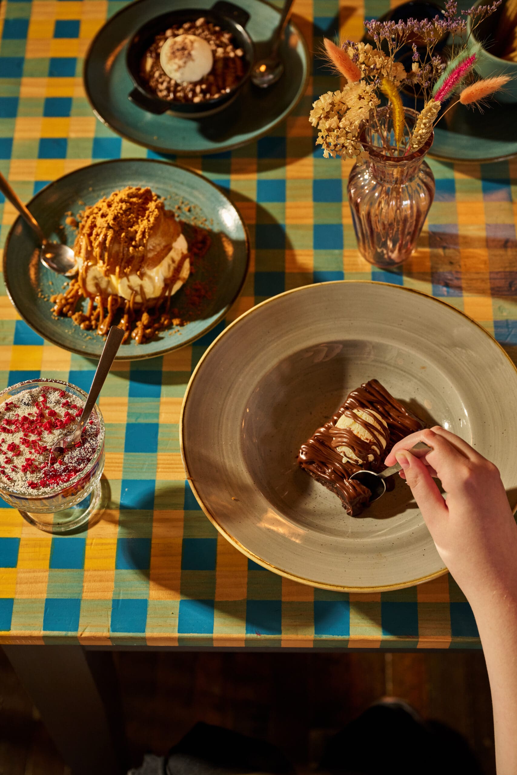 A hand holding a spoon is about to eat a chocolate dessert on a beige plate, surrounded by other desserts, a drink, and a vase with dried flowers on a yellow and blue checkered tablecloth.