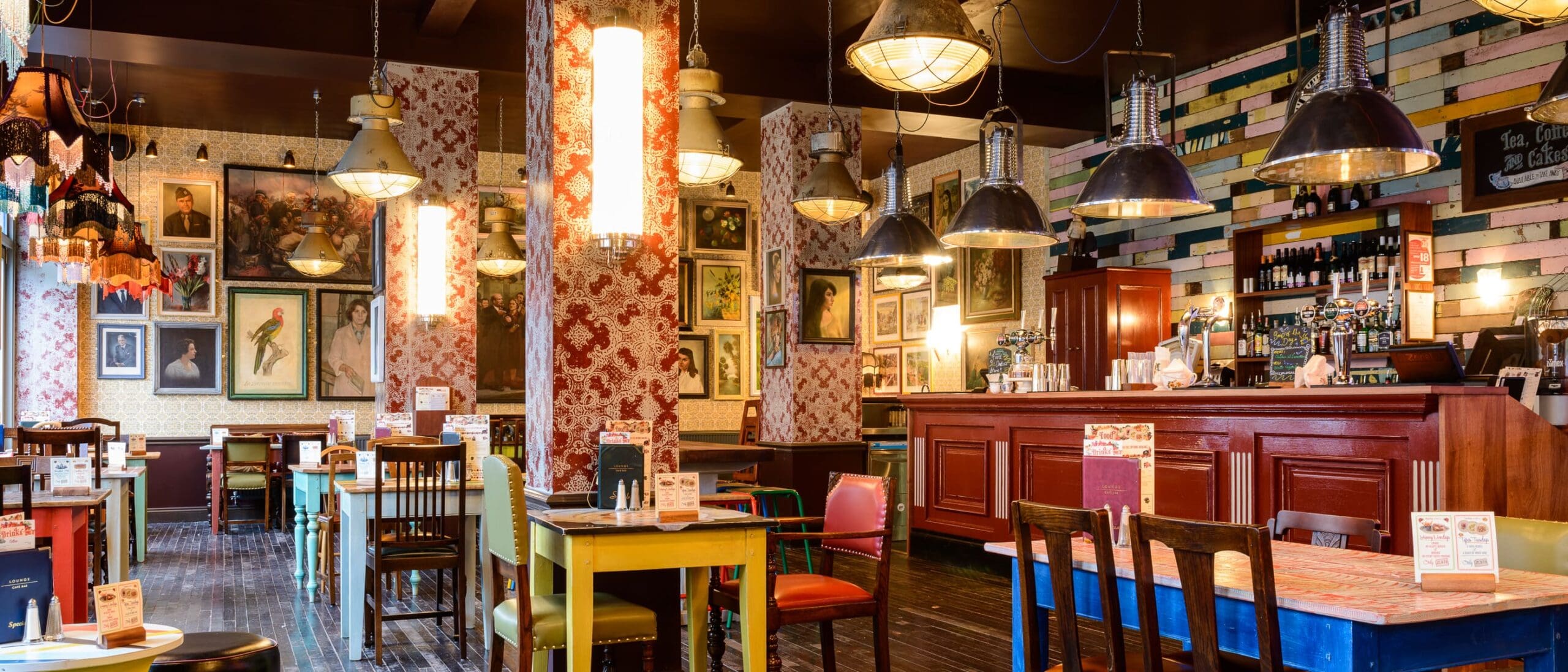 A cozy, eclectic restaurant interior with colorful mismatched chairs and tables, vintage wallpaper, framed artwork, unique hanging lamps, and a wooden bar with Quinto wine bottles and glasses on shelves.