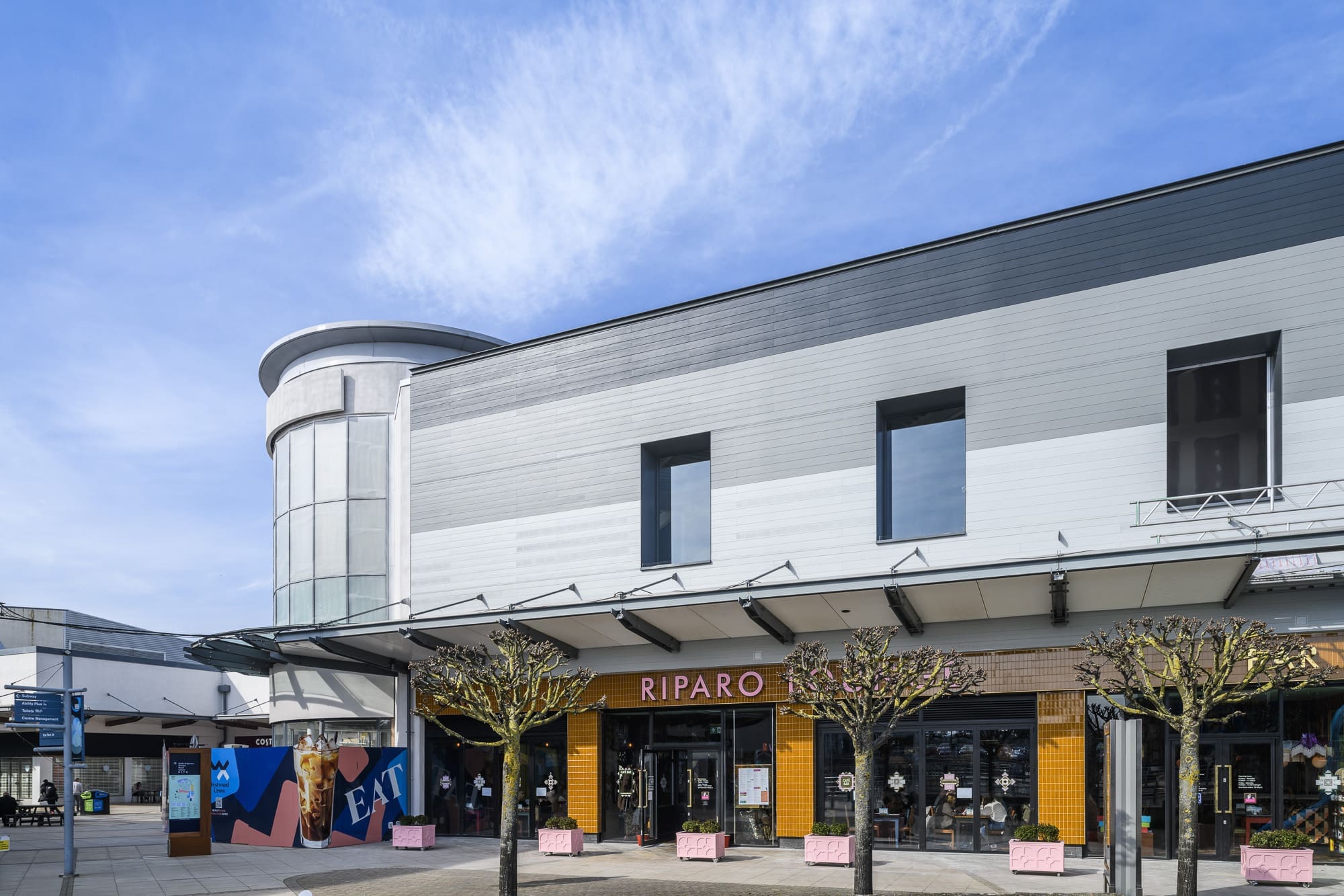 A modern two-story building features the riparo store at street level, with large windows, minimalist design, trimmed trees in planters, and a blue sky overhead.