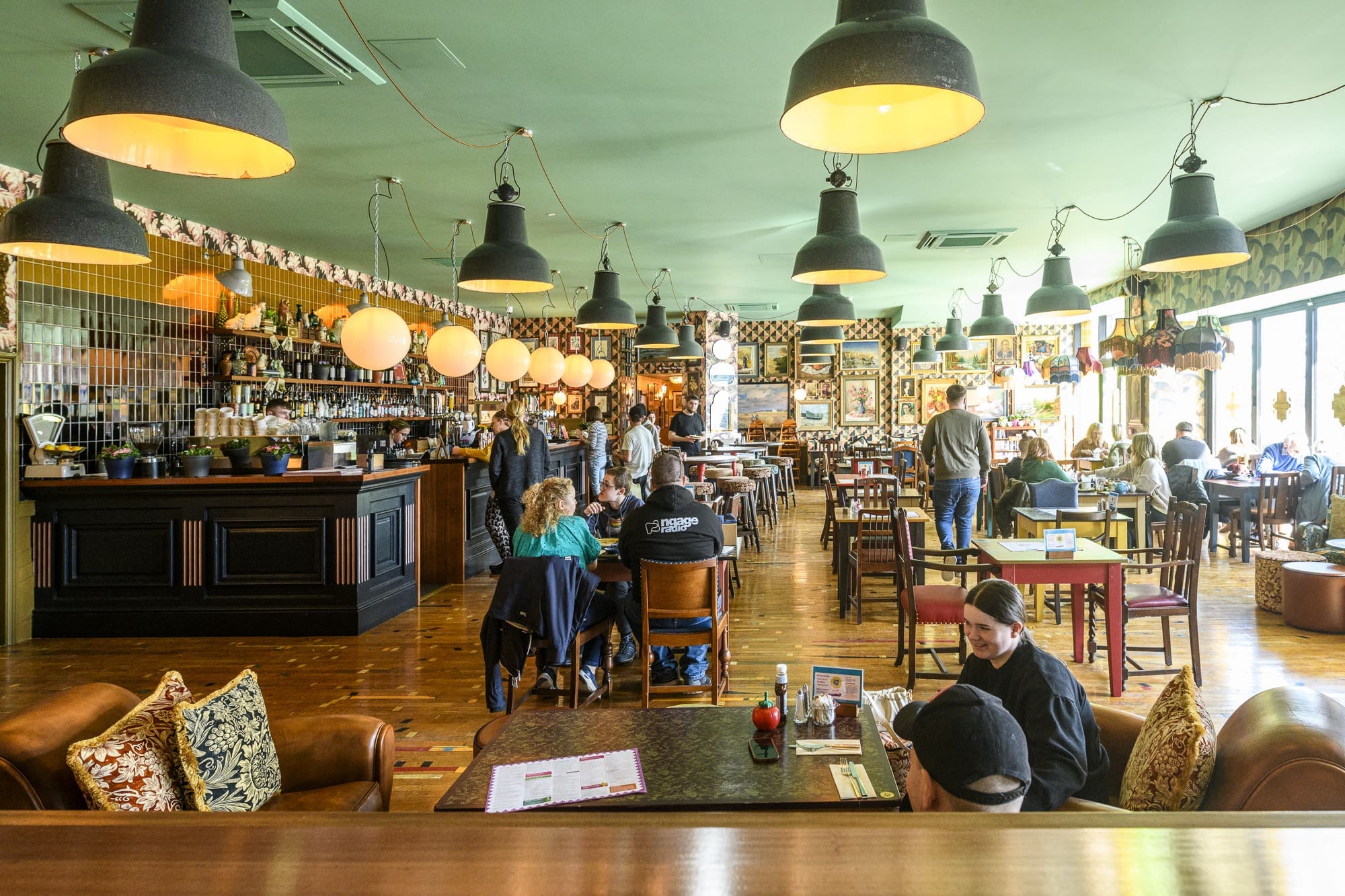 A lively, colorful restaurant interior with people dining at tables, warm hanging lights, and large windows letting in natural light. The bar area acts as a riparo, decorated with plants and bottles to create a cozy, welcoming atmosphere.