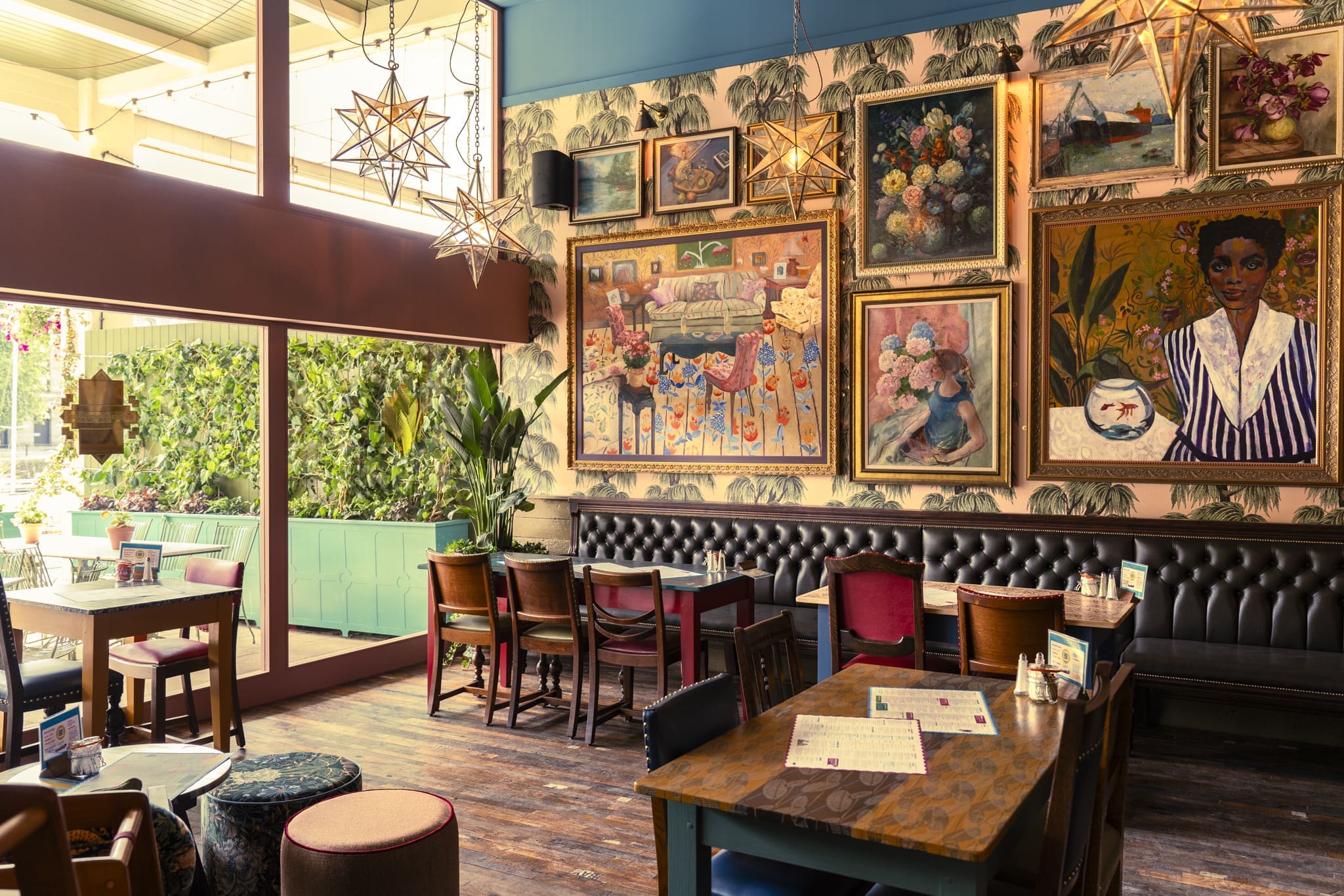 A cozy, eclectic café, Ritorno, with wooden tables and mismatched chairs, features a gallery wall of colorful paintings, patterned wallpaper, and star-shaped pendant lights. Sunlight streams through large windows onto the vibrant space.