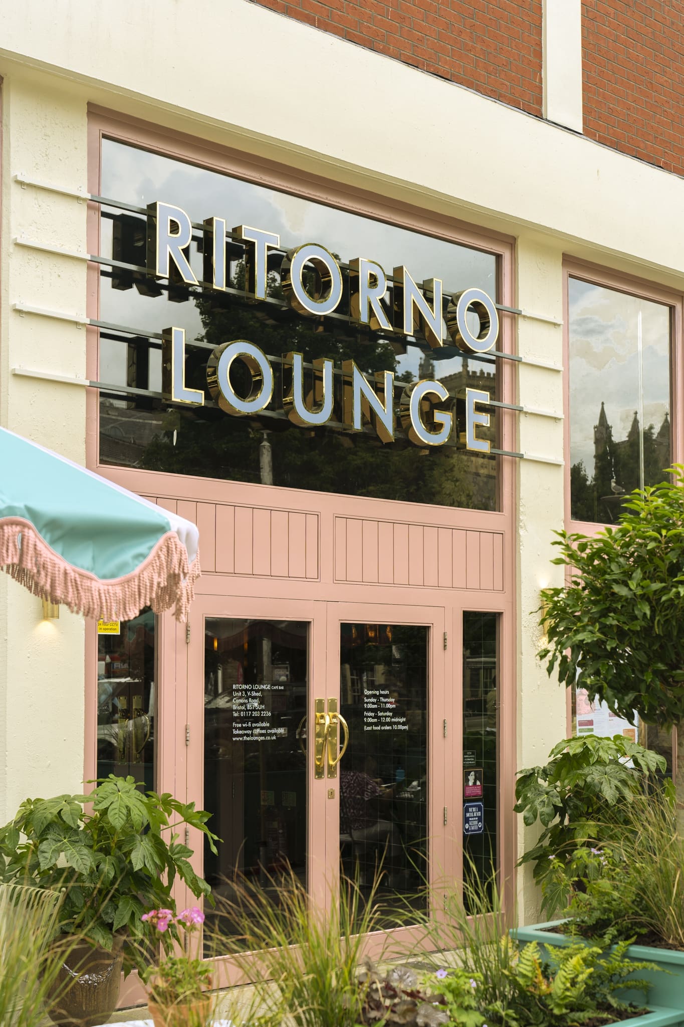 The entrance of Ritorno Lounge features large glass windows, gold Ritorno signage, pink double doors, lush green plants, and part of a pastel umbrella with fringe visible to the left.