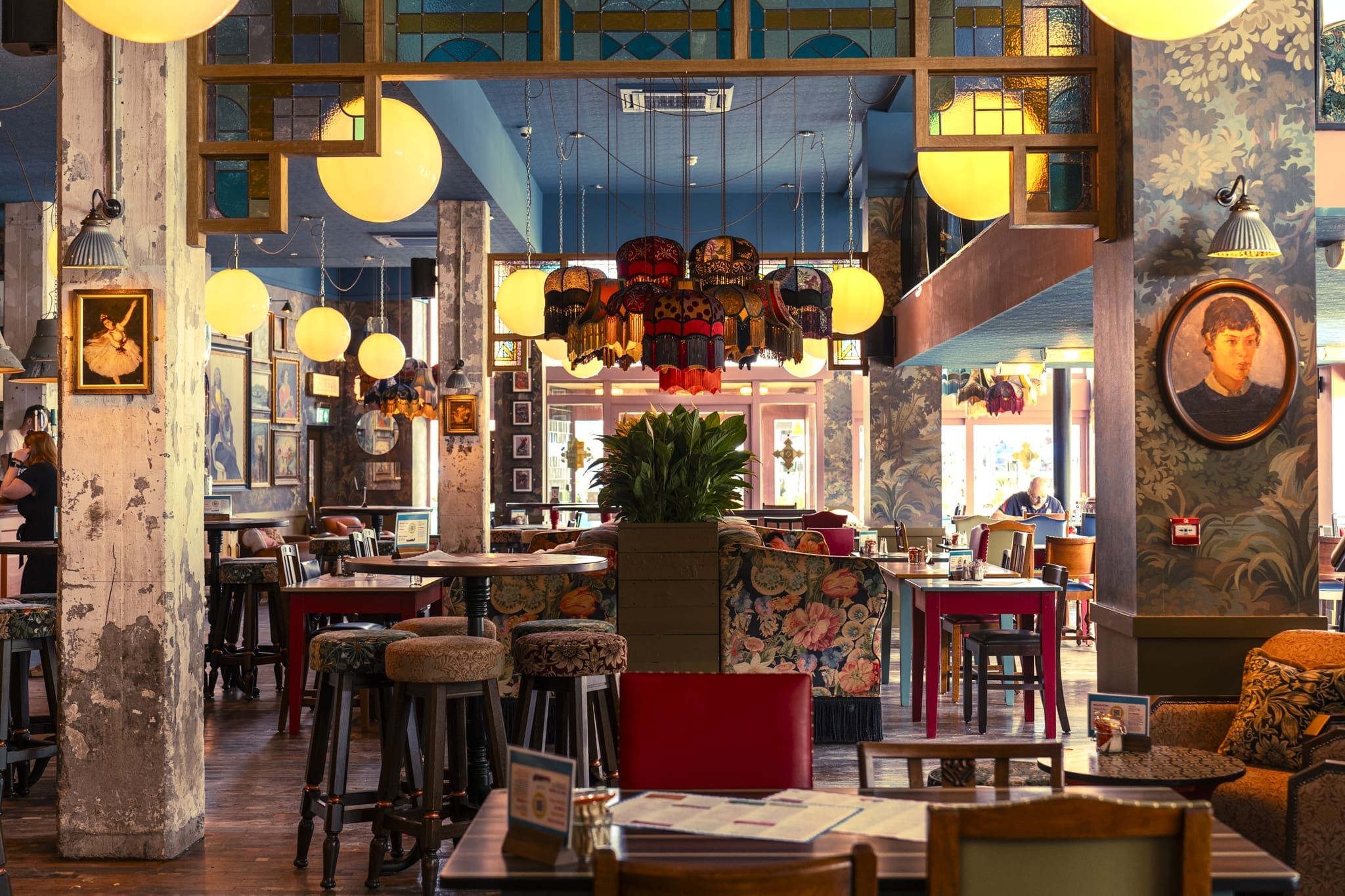 A cozy, eclectic restaurant interior at Ritorno with colorful chairs, patterned wallpaper, round hanging lights, framed portraits, and wooden tables creates a warm and inviting atmosphere.
