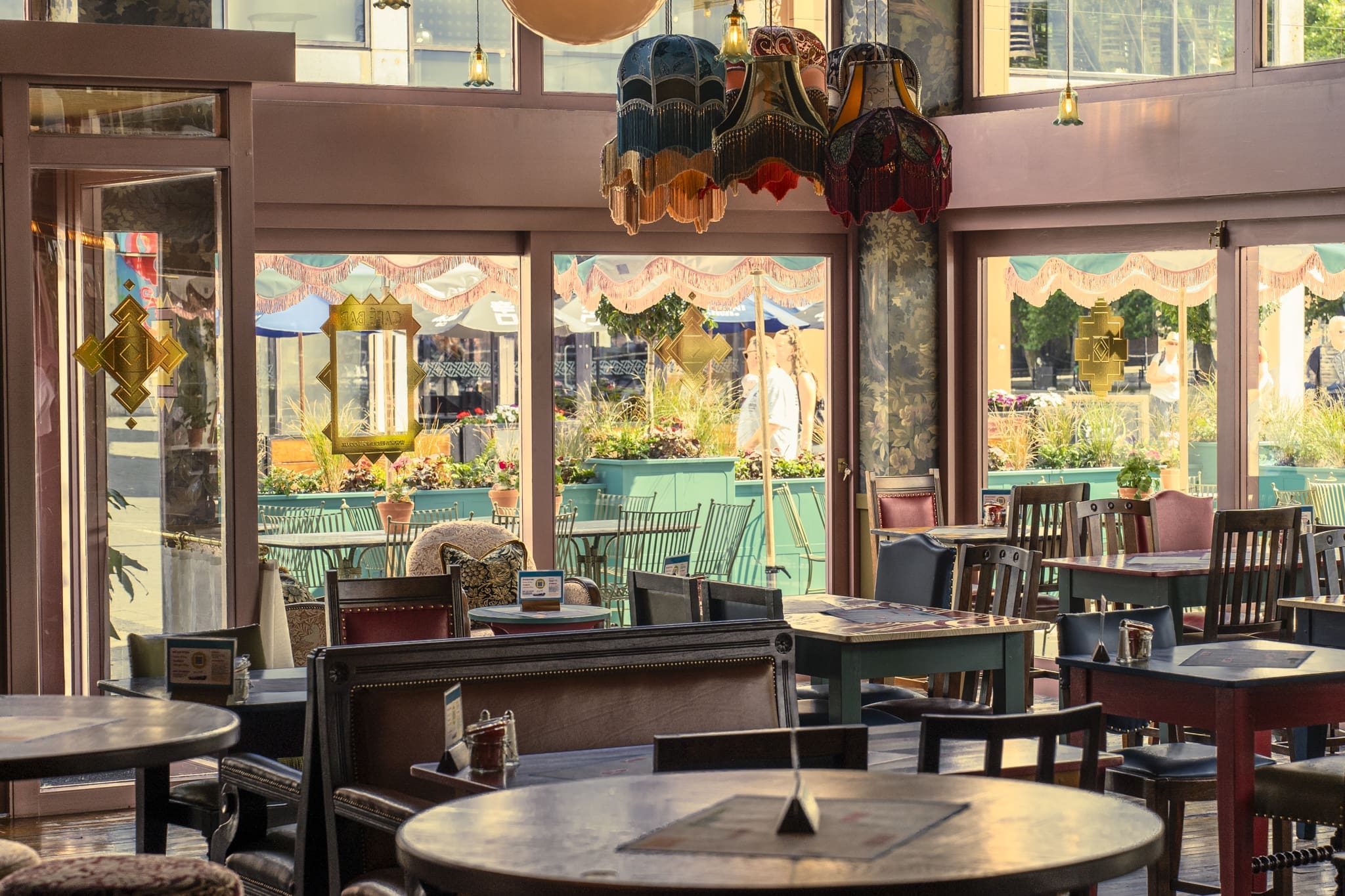 Cozy café interior with mismatched chairs and tables, vintage lamps, and large windows overlooking Ritorno’s outdoor patio with plants and umbrellas. Sunlight streams in, creating a warm and inviting atmosphere.
