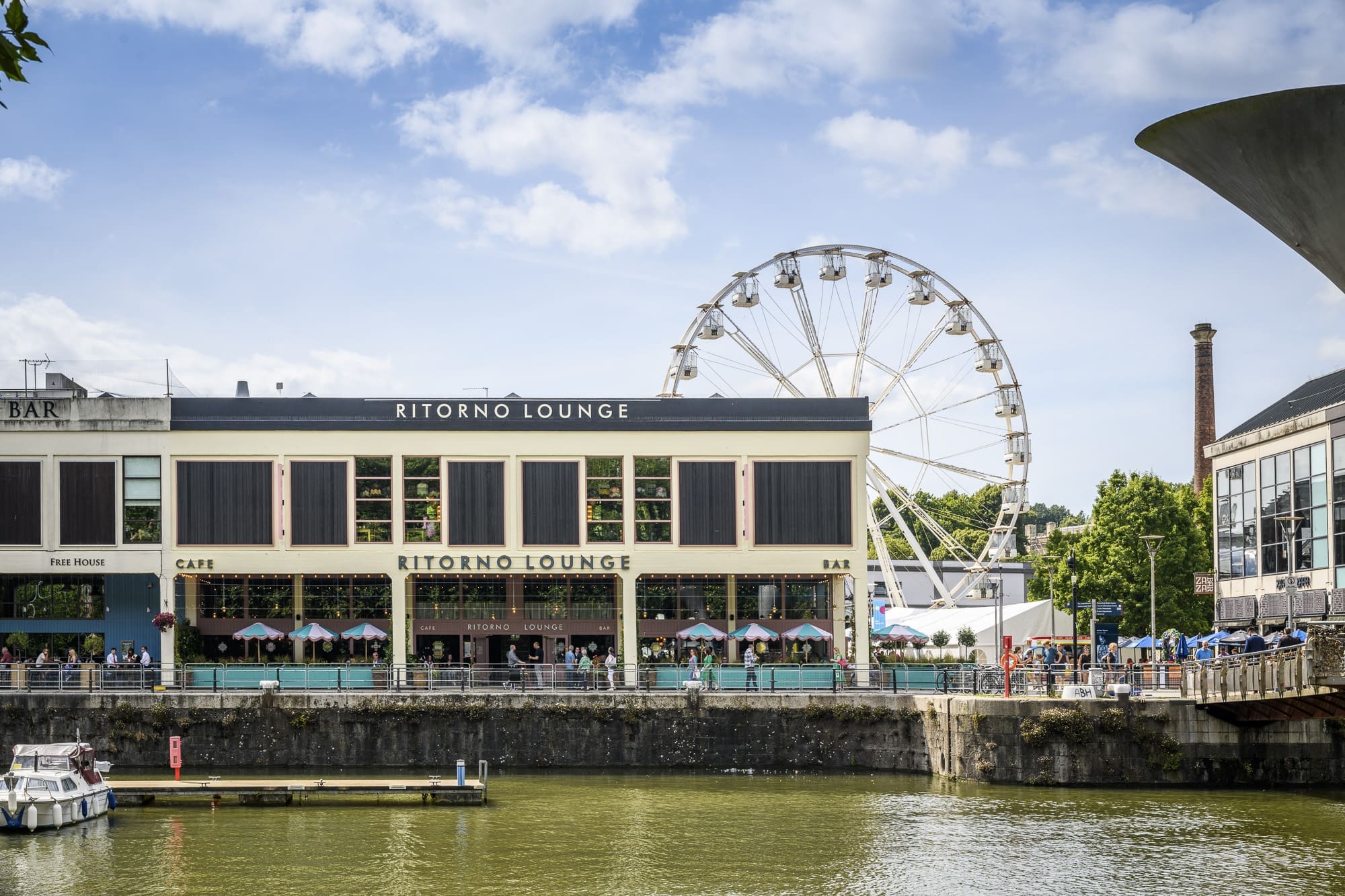 A large riverside building labeled "Ritorno Lounge" has outdoor seating filled with people. A Ferris wheel rises behind it under a blue sky, and the river in the foreground reflects Ritorno and the lively scene.