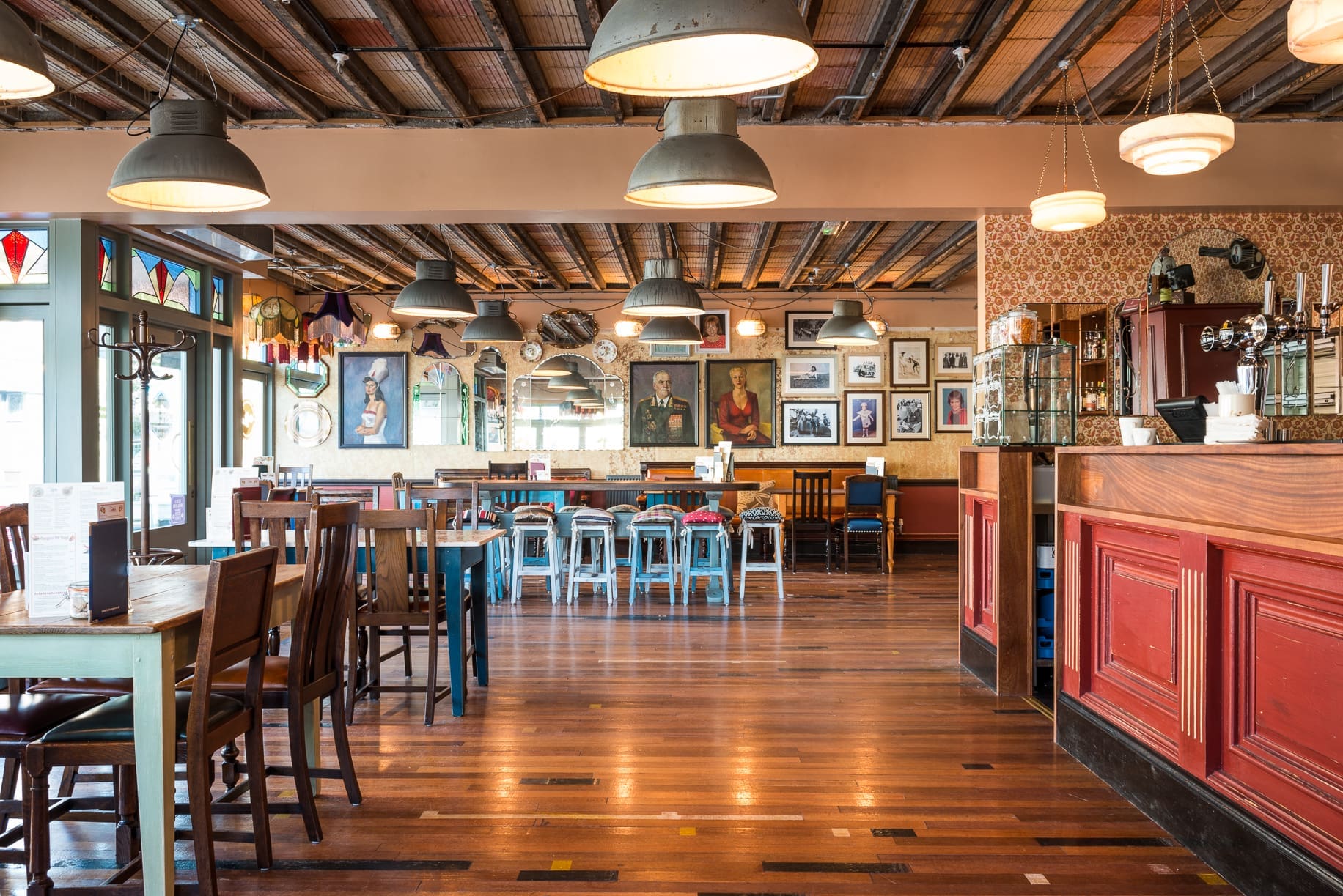 A cozy, vintage-style restaurant, Rivo features wooden floors, mismatched tables and chairs, industrial ceiling lights, and walls adorned with artwork and photos. The red bar counter stands out as natural light streams in through large windows.