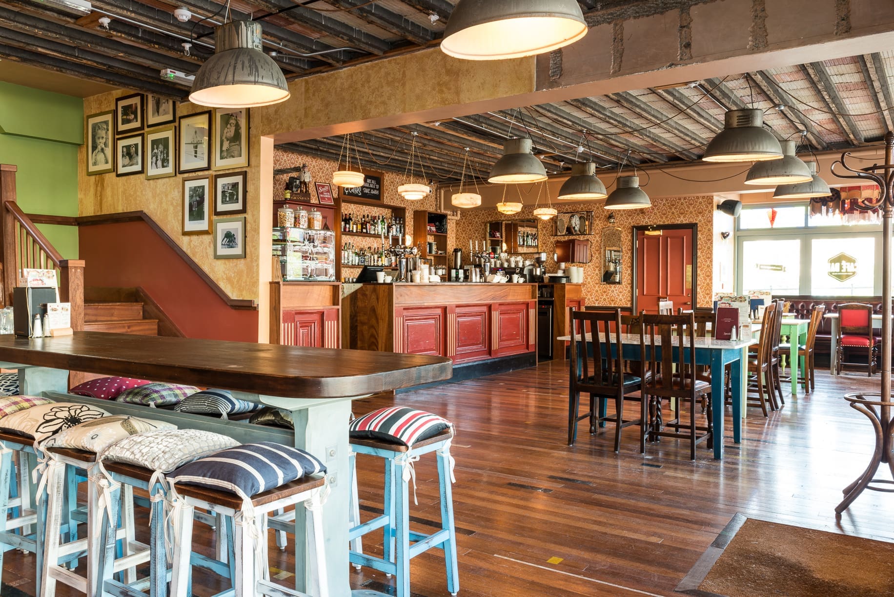 A cozy, well-lit Rivo cafe with wooden floors, eclectic chairs and stools with patterned cushions, a bar counter, framed photos on the wall, and exposed ceiling beams.